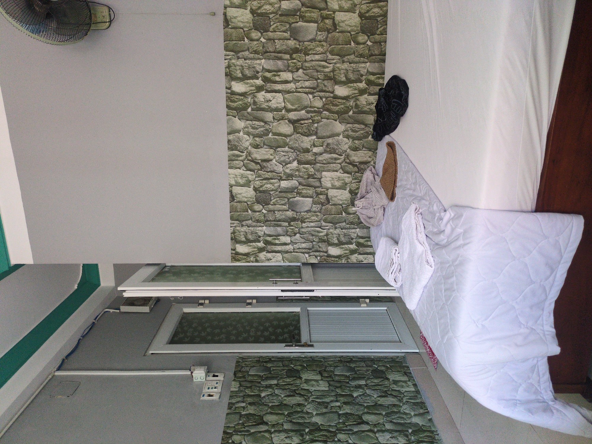 This image depicts an interior room, located in South Quy Nhon Ward, Vietnam, likely a bedroom. The room features light gray painted upper walls, with the lower sections adorned with green-grey stone-patterned wallpaper. The white ceiling has a distinctive green trim. A large bed with a dark brown frame occupies the foreground, covered with white sheets and a white quilted comforter. On the left side of the bed, two neatly folded white towels are stacked. Adjacent to these, a crumpled light-colored, possibly striped, garment, a brown woven flat object, and a dark-colored patterned garment are loosely placed on the bedding. To the left of the bed, a set of aluminum-framed double doors with patterned glass panels is partially ajar, revealing a tiled floor with a small red patterned mat directly in front of the doorway. Electrical conduit, switches, and outlets are mounted on the left wall. A wall-mounted oscillating fan with a dark protective cage and a pull cord is visible on the upper right wall. No individuals are present in the scene.