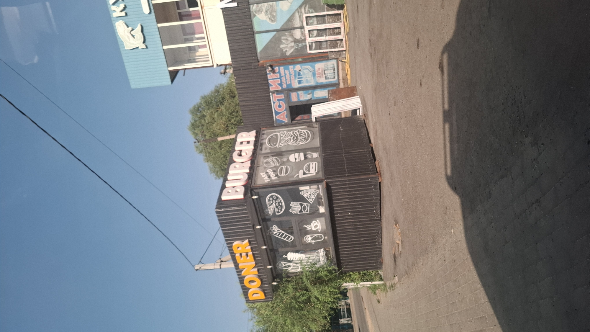 The image captures a street scene in Shubarssu, Kazakhstan, on a clear, sunny day. The focal point is a small food stall with prominent signs reading "DONER" and "BURGER" in large, orange and white lettering. The stall is constructed from corrugated metal and features large glass windows displaying various food illustrations, including pizzas, burgers, French fries, and a doner kebab. A smaller sign inside indicates "FAST FOOD". Adjacent to the food stall, another establishment with a blue facade and a sign with Cyrillic lettering is visible. The ground is paved with interlocking bricks in the foreground, transitioning to asphalt. A shadow of a vehicle falls across the paved area, indicating the presence of a parked car or truck. The environment suggests an urban or suburban setting with a clear blue sky overhead.