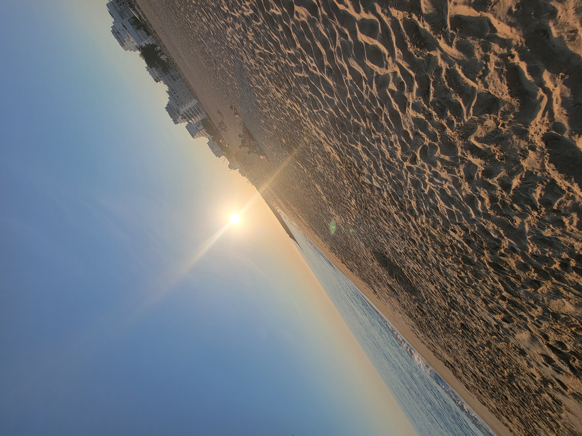 The image captures a sun-drenched beach in Quarteira, Portugal, with the sun low in the sky, casting long shadows and a warm glow. The camera is tilted, giving a dynamic perspective of the scene. The foreground is dominated by textured sand, showing the subtle undulations left by footprints and the tide.
To the left, the calm, blue sea meets the shore with gentle waves. Further inland, a stretch of sandy beach is dotted with a few sunbathers and groups of people, some appearing to be relaxing or walking. Beyond the beach, a line of white, multi-story buildings, characteristic of a coastal resort, stands against the clear blue sky, with trees interspersed between them. The overall impression is one of a peaceful late afternoon or early evening on a popular Portuguese beach.