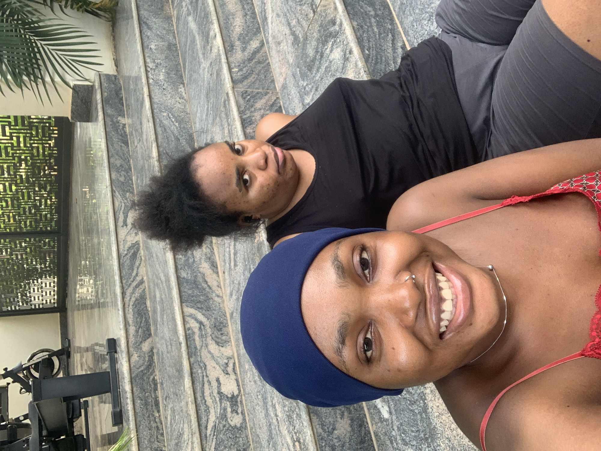 Two individuals are seated on marble steps. The person in the foreground is wearing a navy blue head covering and a red strap top. They have a nose ring and a silver necklace. Their gaze is directed towards the camera with a wide smile. To their right, another individual is wearing a black tank top and dark gray shorts. This person is looking slightly off to the side with a neutral expression. Behind them, there is a decorative metal screen and lush green foliage, suggesting an outdoor setting. In the background, a piece of exercise equipment, possibly a stationary bicycle, is partially visible. The steps are made of polished marble with distinct grey and white veining. The overall impression is of a casual outdoor gathering in a well-maintained environment.