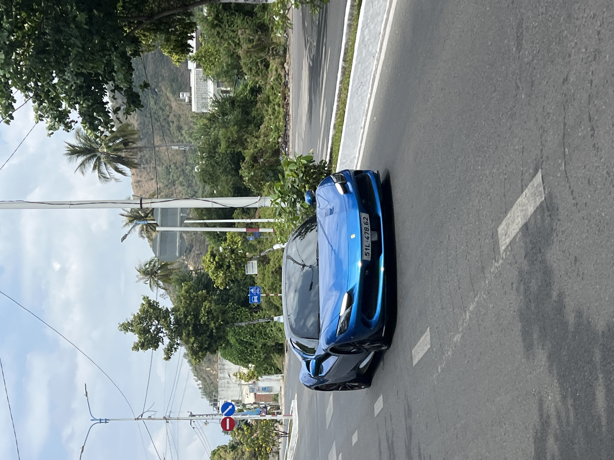 A blue Ferrari, displaying a license plate that reads "51L-478.62", sits on a paved road in what appears to be the city of Bắc Nha Trang, Vietnam. The car is angled diagonally in the frame, with the front of the vehicle facing towards the upper right corner. A white curb is visible alongside the road. Lush greenery, including trees and palm trees, lines the side of the road. Buildings and a mountain are visible in the background. Power lines crisscross the sky. Road signs are posted near the left side of the road. The sky is partly cloudy.