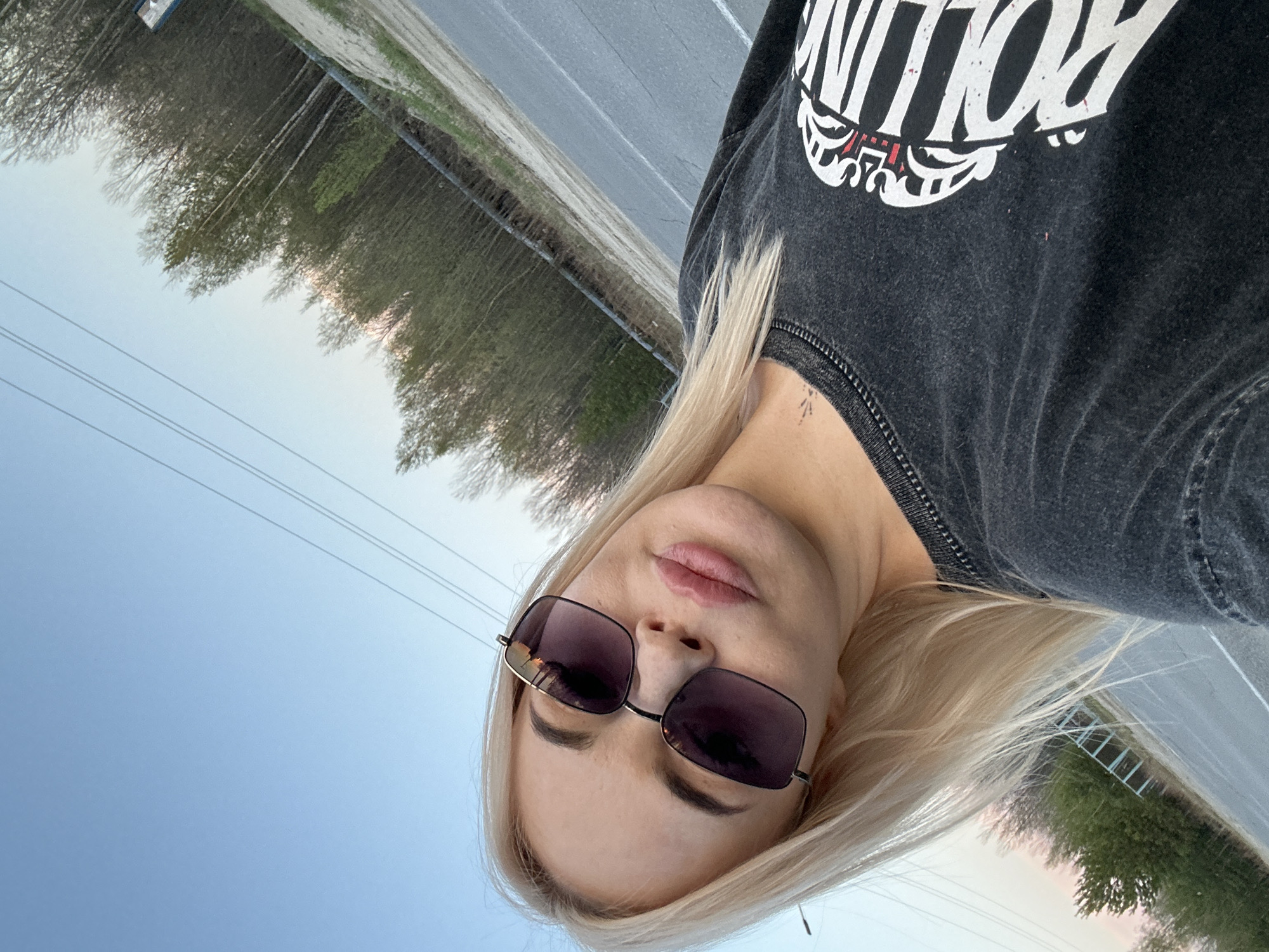 The photograph is taken from a low angle, showcasing a woman with blond hair wearing sunglasses and a dark gray sweatshirt with white writing. A small tattoo is visible on her neck. In the background, a road stretches towards the horizon, flanked by trees. The sky is a pale blue, suggesting early morning or late evening light. Based on the surrounding environment, the location may be outside of the city proper on one of the highways in the area. The road is paved and in good condition. The woman is looking directly into the camera.