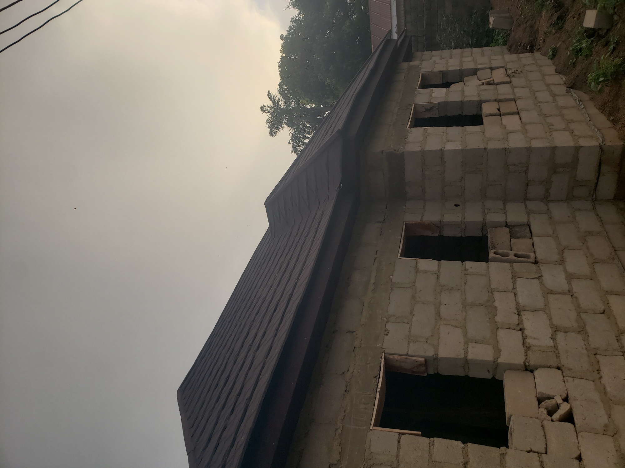 The image shows a partially constructed building in Bukuru, Nigeria. The building is made of concrete blocks and has a brown roof. There are several window openings, some of which are filled with broken blocks. The surrounding environment includes vegetation and trees. The sky is overcast. There are power lines visible in the upper left corner of the image.