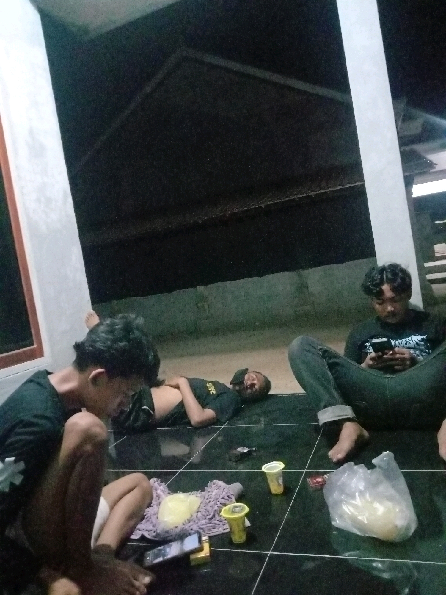 Three young men are gathered on a tiled floor at night, possibly in a covered outdoor area. The scene suggests a casual social gathering. One man, in the foreground and to the left, is sitting on the floor and appears to be interacting with his phone, which is placed on the floor in front of him. His legs are bent, and he is wearing a dark t-shirt. To his right, another man is lying on his back on the floor, with his head resting on his arm and his eyes closed, appearing to be asleep or resting. He is wearing a dark t-shirt with some white lettering. A third man is seated to the right, looking down at his phone, which he is holding in his hands. He is wearing a dark t-shirt and blue jeans. Scattered on the floor are two small yellow cups, possibly containing drinks or snacks, a plastic bag filled with a light-colored substance, a purple mat, and some small items that are difficult to identify. The background shows a dark, textured ceiling and a concrete wall. The lighting is dim, indicating it is nighttime. The location in Utama Jaya Mataram, Indonesia, is an assumption based on the request and cannot be definitively determined from the image alone.