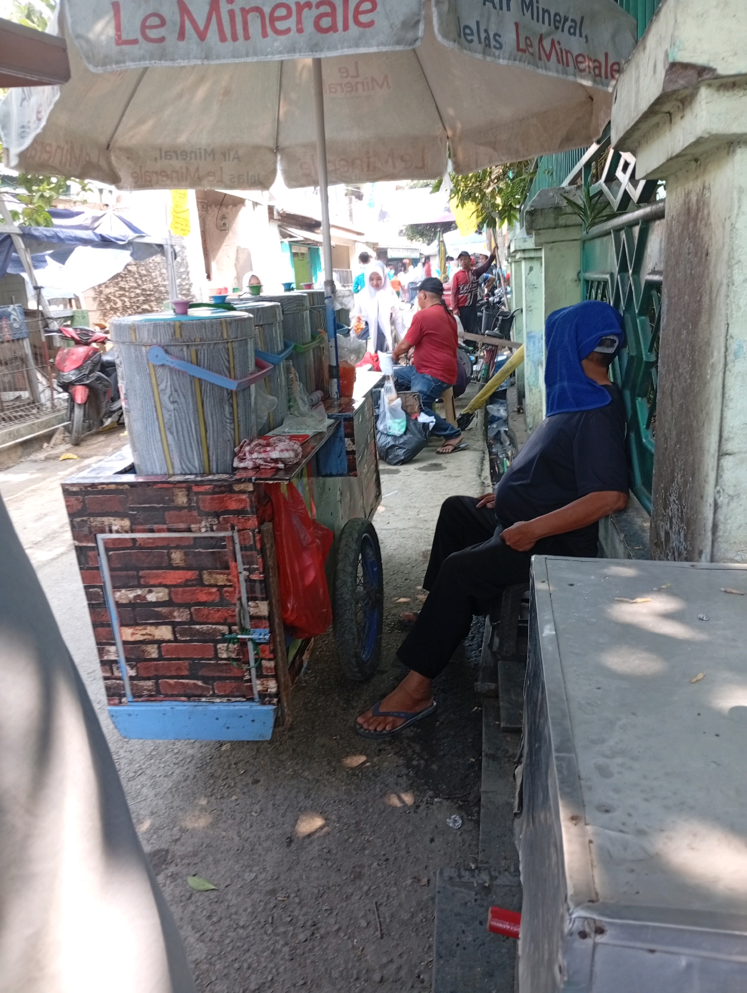 In a city street setting, likely in East Jakarta, Indonesia, a man wearing a blue headscarf and sunglasses sits with his back against a weathered concrete wall and a green metal fence. He is dressed in a black t-shirt and black trousers, with his right hand resting on his lap. The man appears to be resting or sleeping, with the headscarf obscuring his face. To his left, a mobile food cart is parked. The cart is decorated with a brick pattern on its front and sides. On top of the cart are several large, cylindrical containers, possibly for drinks, with blue and red accents. A red plastic bag hangs from the cart, and a spare tire is attached to its side. In the background, other people and motorcycles are visible, suggesting a busy street environment. A large, faded umbrella, advertising "Le Minerale Air Mineral," is positioned above the cart, providing shade.
