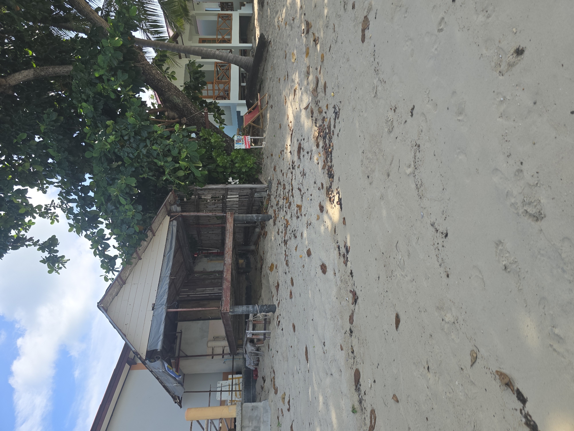 The image depicts a beach scene in Baan Lamai, Thailand, characterized by sand, a partly constructed wooden structure, and a modern building in the background. The foreground is dominated by light-colored sand with scattered dry leaves and footprints. To the left, a partially covered wooden structure on stilts is visible, with exposed wooden beams and supports. Behind this structure, a white building with an orange-tiled roof is partially visible. To the right, a contemporary white building with wooden balconies and railings is situated, with a large tree in front of it, casting shadows on the sand. A red lounge chair and a small sign are placed on the sand near the building. The sky above is blue with scattered white clouds.