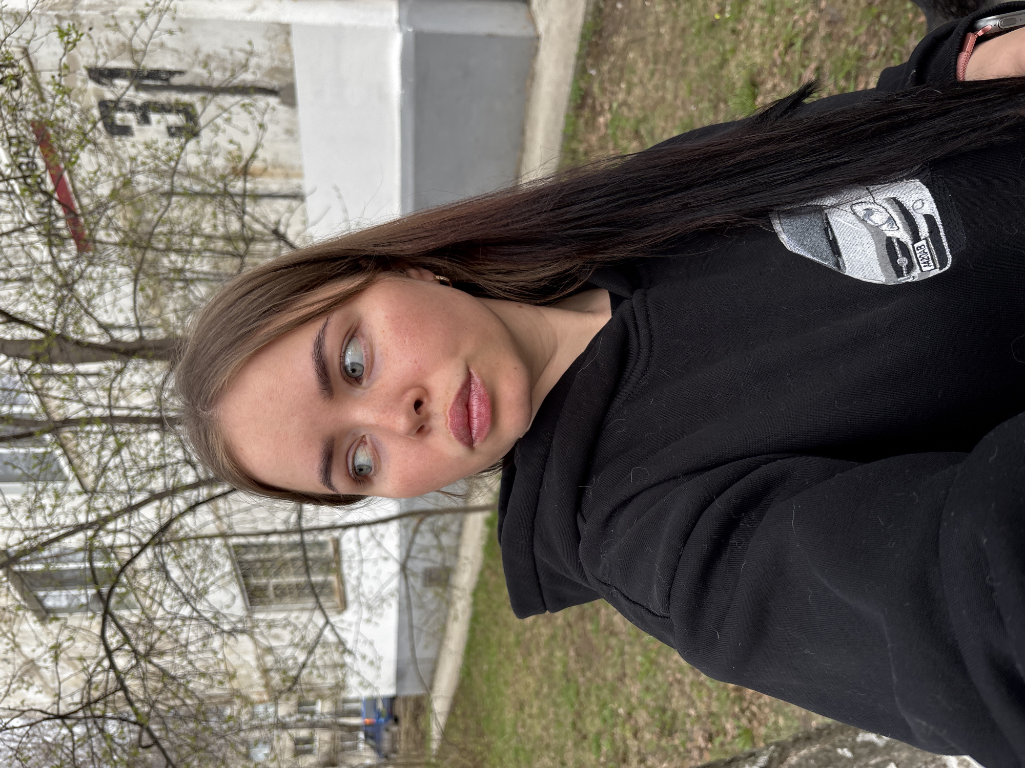 A young woman, likely in her late teens or early twenties, is depicted in an outdoor setting, appearing to take a selfie. She has long, straight dark brown hair, fair skin, and light-colored eyes (appearing greenish-grey). Her lips are slightly parted, and she has a wide-eyed, somewhat surprised or curious expression. She is wearing a black hooded sweatshirt (hoodie) with small fibers/lint visible on the fabric. A small gold-colored stud earring is visible in her right ear, and a fitness tracker or digital watch with a pink strap can be seen on her left wrist, partially covered by her hair.
An embroidered patch depicting the rear-left view of a car, resembling a first-generation Mazda MX-5 (NA), is visible on the left shoulder/upper arm of her hoodie. The car's license plate in the embroidery clearly reads "MAZDA."
The background suggests an urban or suburban environment, typical of Izhevsk, Russia. Behind her, light-colored, multi-story residential buildings are visible, with dark grey trim along their bases. Bare tree branches with newly sprouting green leaves indicate early spring. Green grass covers the ground in the mid-ground. The lighting is soft and diffused, suggesting an overcast daytime.
On a distant building, partially obscured by tree branches, blurry black characters resembling the Cyrillic letters "Э3" are visible against a white wall.