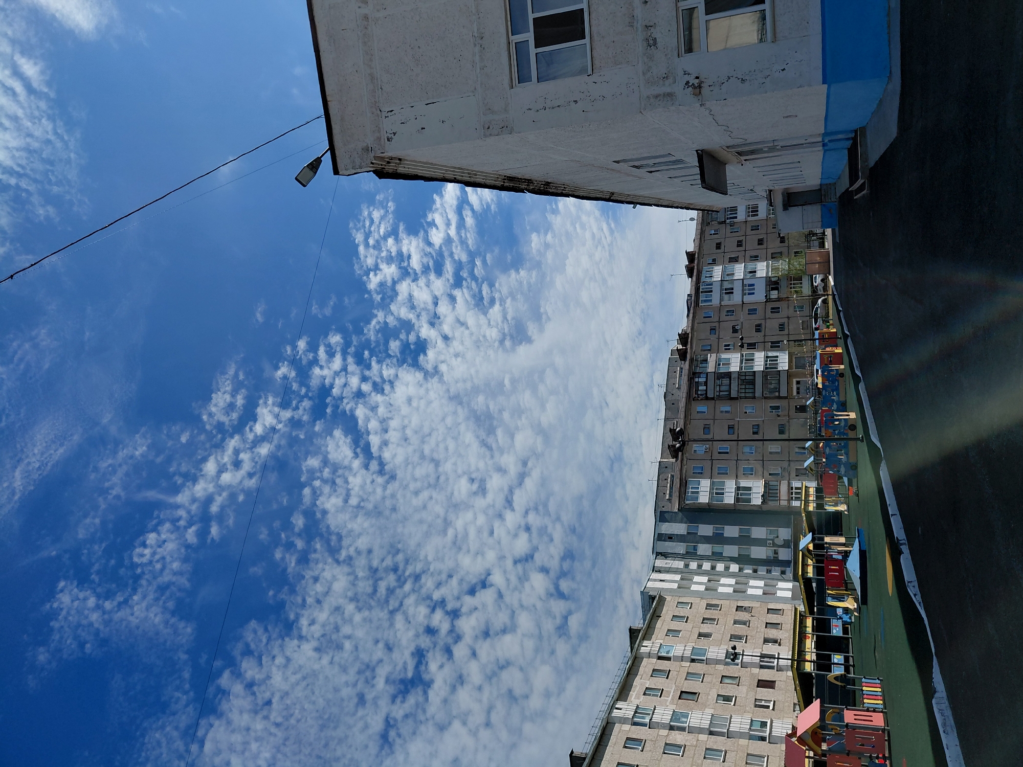 This image presents a skewed, upward-looking perspective of a residential area in Nadym, Russia, under a bright blue sky. The sky is adorned with numerous small, scattered cumulus clouds, creating a textured "mackerel sky" appearance.
The scene is dominated by several multi-story apartment buildings, characteristic of Soviet-era or post-Soviet architecture. In the immediate foreground, an older building with a weathered, light-colored facade features a horizontal blue stripe and multiple windows. A street light and power lines stretch across the clear sky from this building. Further in the background, a row of other apartment blocks, primarily grey and beige, with many windows, extends across the frame.
At the bottom right, a dark, paved surface, likely a sidewalk or playground area, is visible, marked by a white line. Beyond this, colorful structures of a children's playground can be seen. The time of day is clearly daylight, under sunny conditions. There is no visible human activity or discernible text in the image. The overall impression is a quiet, bright day in an urban residential district.