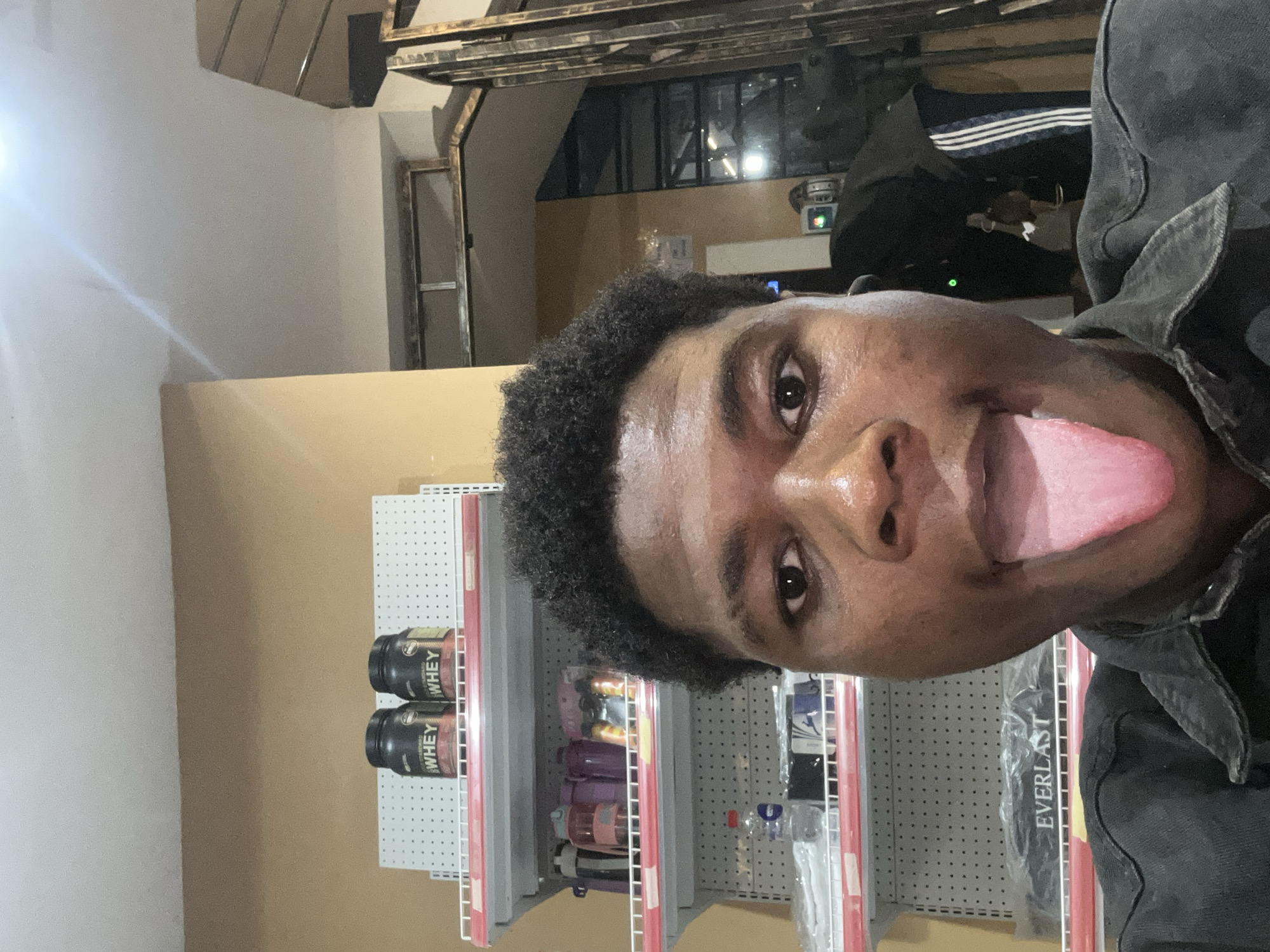 A medium shot captures a young Black person with curly hair and brown eyes, sticking out their tongue. They are wearing a dark denim jacket. The background features shelves stocked with various items, including two large tubs labeled "WHEY," several water bottles, and a packaged item with "EVERLAST" printed on it. The environment appears to be a store or a similar retail space. The lighting suggests an indoor setting, potentially with artificial lights contributing to the overall illumination. The background also includes a staircase and a wall with a yellow-beige hue.