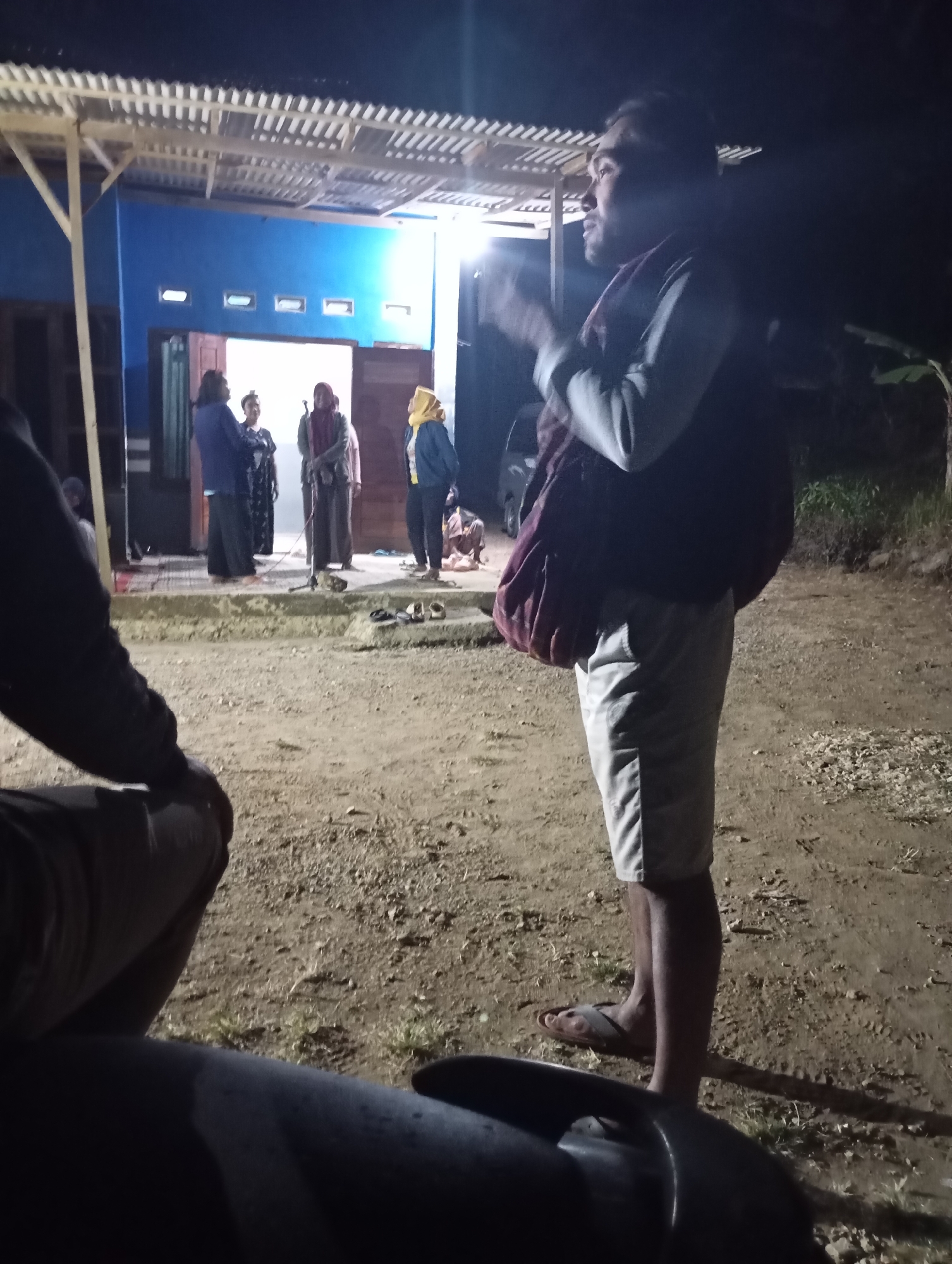 The image depicts an outdoor gathering at night in the city of Ketepung, Indonesia. In the foreground, a person is partially visible, their body angled towards the right, seated on or near a dark, metallic object, possibly a motorcycle. Behind them, the ground is rough and sandy, with sparse vegetation.
Further in the background, a group of individuals stands on an elevated platform in front of a blue building with a white, slatted roof structure. The building appears to be a residential or community structure, with doorways and windows visible. Several people are gathered on the platform, dressed in traditional clothing, including headscarves. One person is holding a microphone stand. The scene is illuminated by artificial lighting, casting shadows and creating a somewhat dim atmosphere. The overall impression is of a casual nighttime social or community event.