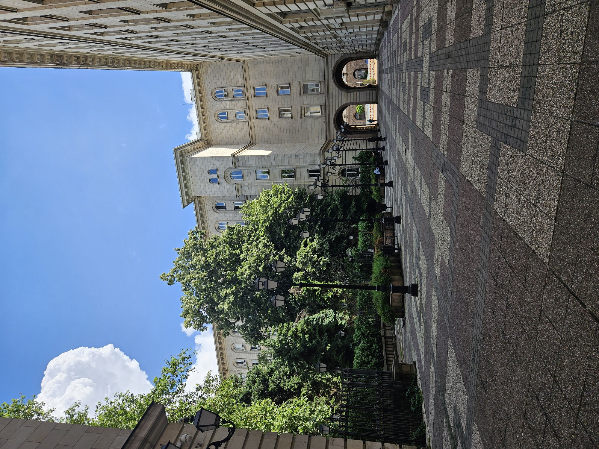 This media file captures a sunny daytime view of a grand architectural passage or courtyard in Sofia, Bulgaria. The scene is dominated by two large, multi-story buildings of classical design, constructed from light-colored stone, which flank a patterned paved pathway. The buildings feature numerous arched and rectangular windows, along with detailed cornices and architectural accents.
On the left side of the passage, a dense arrangement of lush green trees and bushes provides a vibrant natural contrast to the urban stonework, with some foliage extending upwards towards the sky. The pathway itself is distinctive, laid with alternating sections of light and dark rectangular tiles, creating a strong geometric pattern that leads the eye into the distance. Along the right side of the path, several ornate black lampposts, some featuring multiple lights, stand sentinel, bordering a fenced garden area.
In the background, multiple archways pierce through the far building, leading to another area beyond and hinting at a larger complex. Through these arches, indistinct figures can be seen, suggesting pedestrian activity. Overhead, a bright blue sky is adorned with large, fluffy white cumulus clouds, indicating clear and pleasant weather. No discernible text is visible. The overall impression is one of historical grandeur and an inviting public space bathed in natural light.
