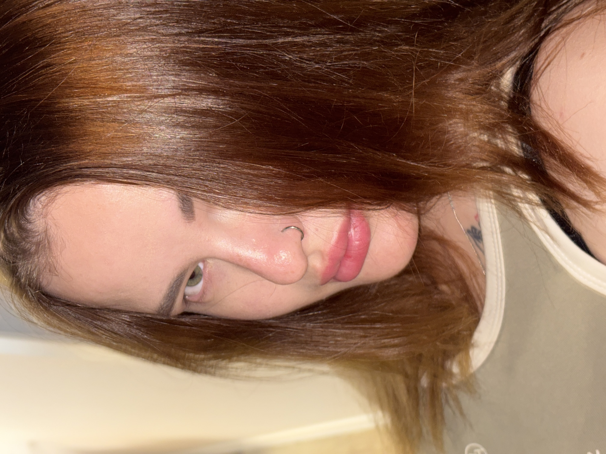 A close-up, angled portrait features a person with reddish-brown/auburn hair, tilted horizontally within the frame. The subject's face occupies the left side of the image, with long, straight hair fanning out towards the right. Her skin appears clear, and she has visible green eyes, dark eyebrows, and plump lips with a pinkish gloss. A silver hoop nose ring is visible on her right nostril.
She is wearing a light-colored, possibly beige or pale green, tank top or camisole, part of which is visible over her right shoulder and chest area. A small, circular, indistinct logo or design can be seen on the fabric near the bottom right. On her neck/shoulder, a faint, dark tattoo is partially visible, appearing to be a floral or abstract design.
The lighting suggests an indoor setting, possibly with artificial light, creating a focused illumination on the subject. The background is a soft, out-of-focus light tone, likely a wall or a simple surface. The person's expression is neutral, with her gaze directed towards the viewer. The overall composition highlights her facial features and hair. No specific location cues are visible within the image itself; however, the location context provided is Krasnodar, Russia.