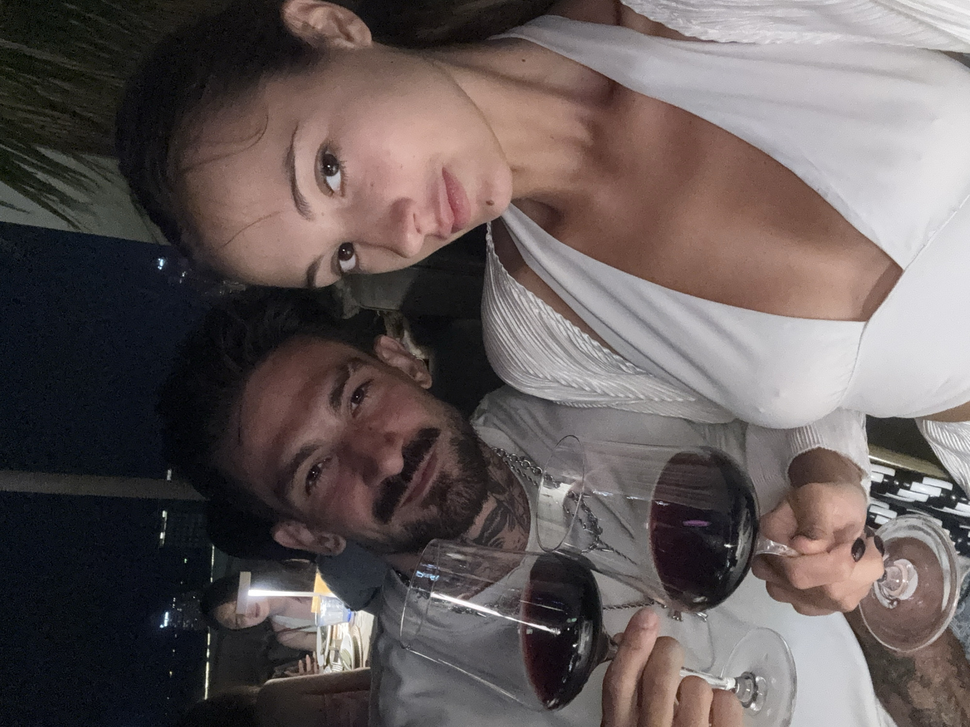 A couple poses for a selfie. The woman, wearing a white top, is positioned above the man, who has a beard and tattoos. They are holding wine glasses filled with red wine. The background is dark, with lights suggesting a nighttime setting in Pattaya City, Thailand. Other people are seated at a table in the background.