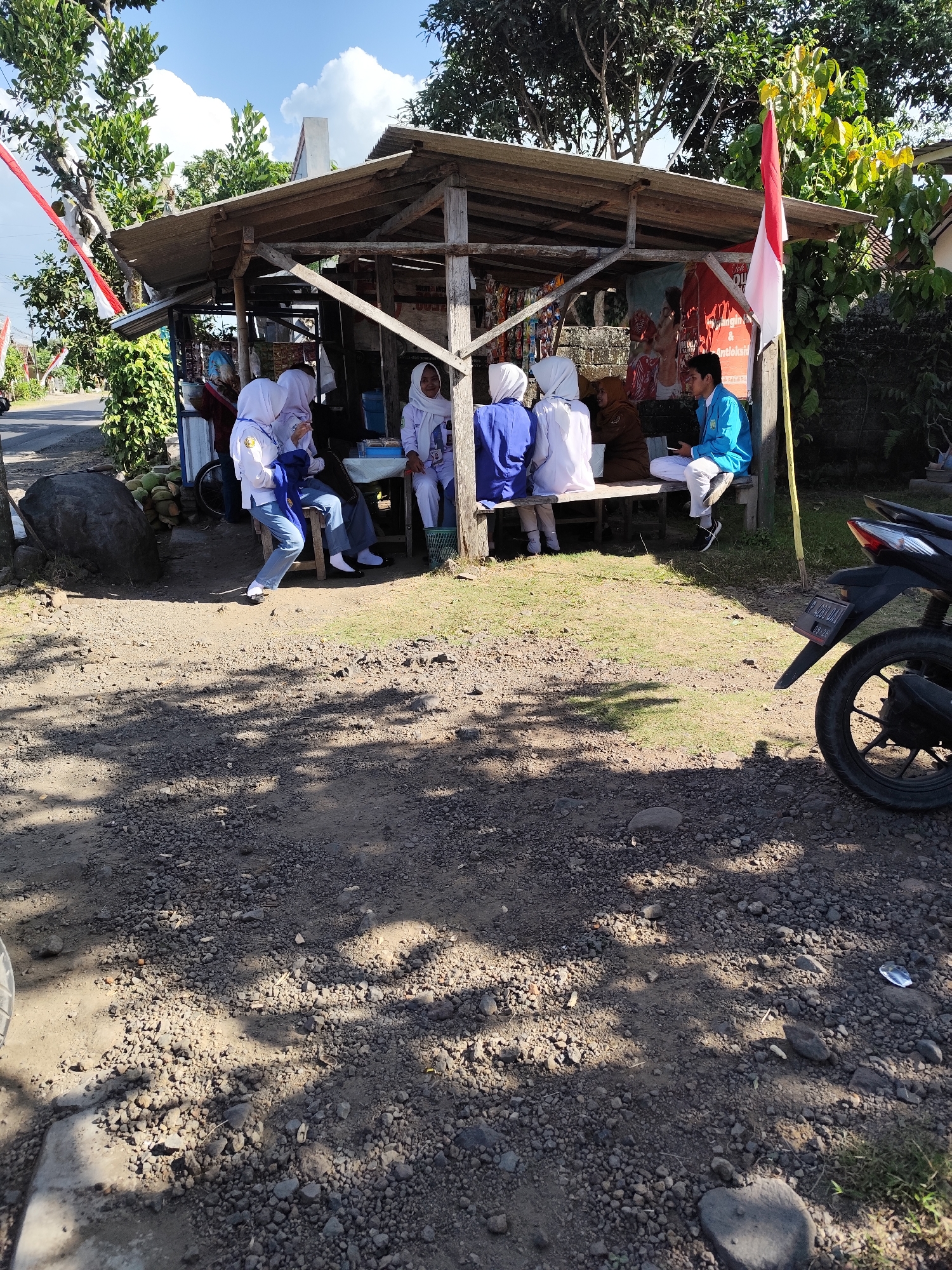 The image depicts an outdoor scene in Tegalarum, Indonesia. In the foreground, the ground is covered with gravel and dirt, with patches of grass. A motorcycle is parked on the right side of the frame. A simple wooden structure, likely a roadside stall, is visible in the middle ground. Several people are gathered under the stall. Four individuals wearing white uniforms with blue accents, possibly students, are seated on a wooden bench. Another person in a blue jacket and white pants is also seated. The stall has a corrugated metal roof and is adorned with Indonesian flags. In the background, lush green trees and vegetation are present, with a bright blue sky with white clouds above. The overall impression is a casual gathering at a local marketplace or rest stop.