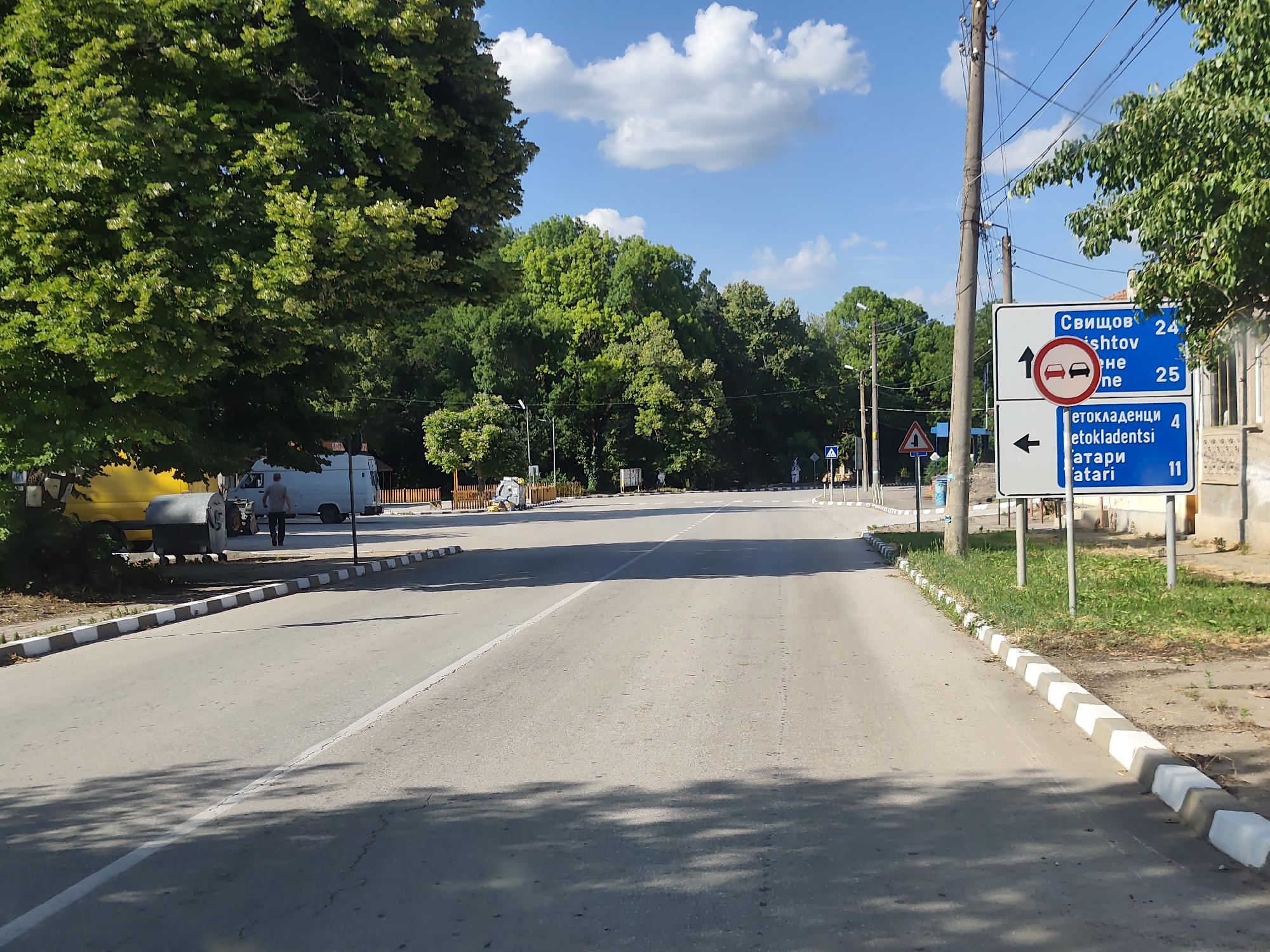 The image shows a street scene in Strezherovo, Bulgaria, on a sunny day with scattered clouds. The road is paved and has a white line marking the lanes. On the right side of the road, there is a traffic sign with destinations and distances written in Bulgarian. The sign indicates Svishtov is 24 km away and another destination is 25 km away. Below that, it shows a destination is 4 km away to the left, and another is 11 km away. There are also trees and vegetation on both sides of the road. A person is walking away from the camera on the left side of the road, near a yellow van and a white utility vehicle. In the background, there are more trees and buildings.