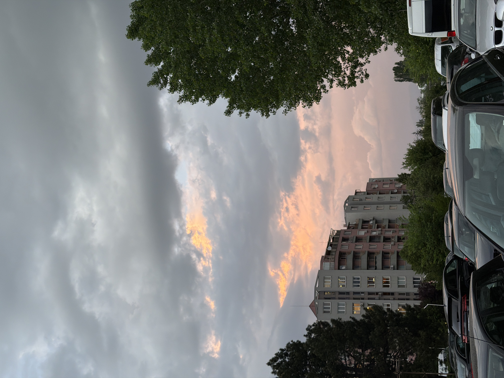 The image shows an outdoor scene in Sofia, Bulgaria, likely a parking lot adjacent to an apartment building. The sky is predominantly cloudy with grey and pink hues, possibly during sunset or sunrise. A multi-story apartment building is visible, featuring balconies and a red tiled roof. Several parked cars are in the foreground, some with doors open. Trees frame the view at the top and bottom of the image. There are no visible people.