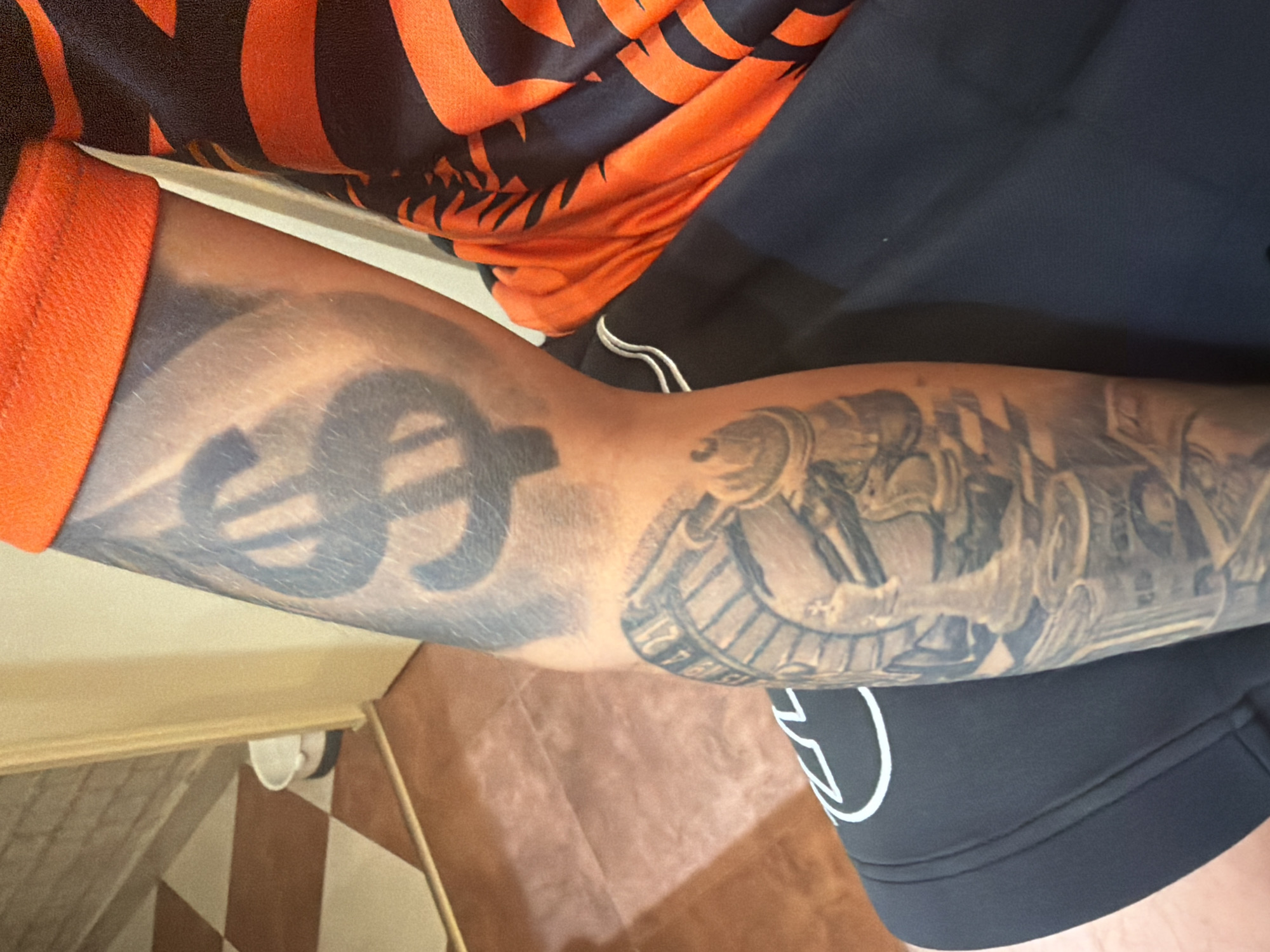 A person's forearm, covered in a tattoo of a dollar sign and a roulette wheel, is visible. The person is wearing an orange and black striped shirt and black sweatpants. The background shows a tiled floor and a doorway.