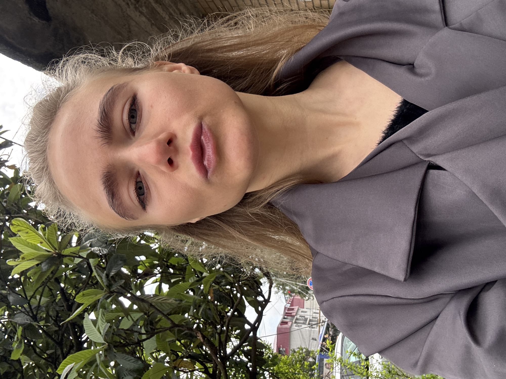The image shows a young woman with blonde hair wearing a grey jacket. She is outdoors, with a lush green tree with large leaves visible in the lower left corner. Buildings can be seen in the background. The location appears to be an urban area with trees and foliage. The exact location is unknown.