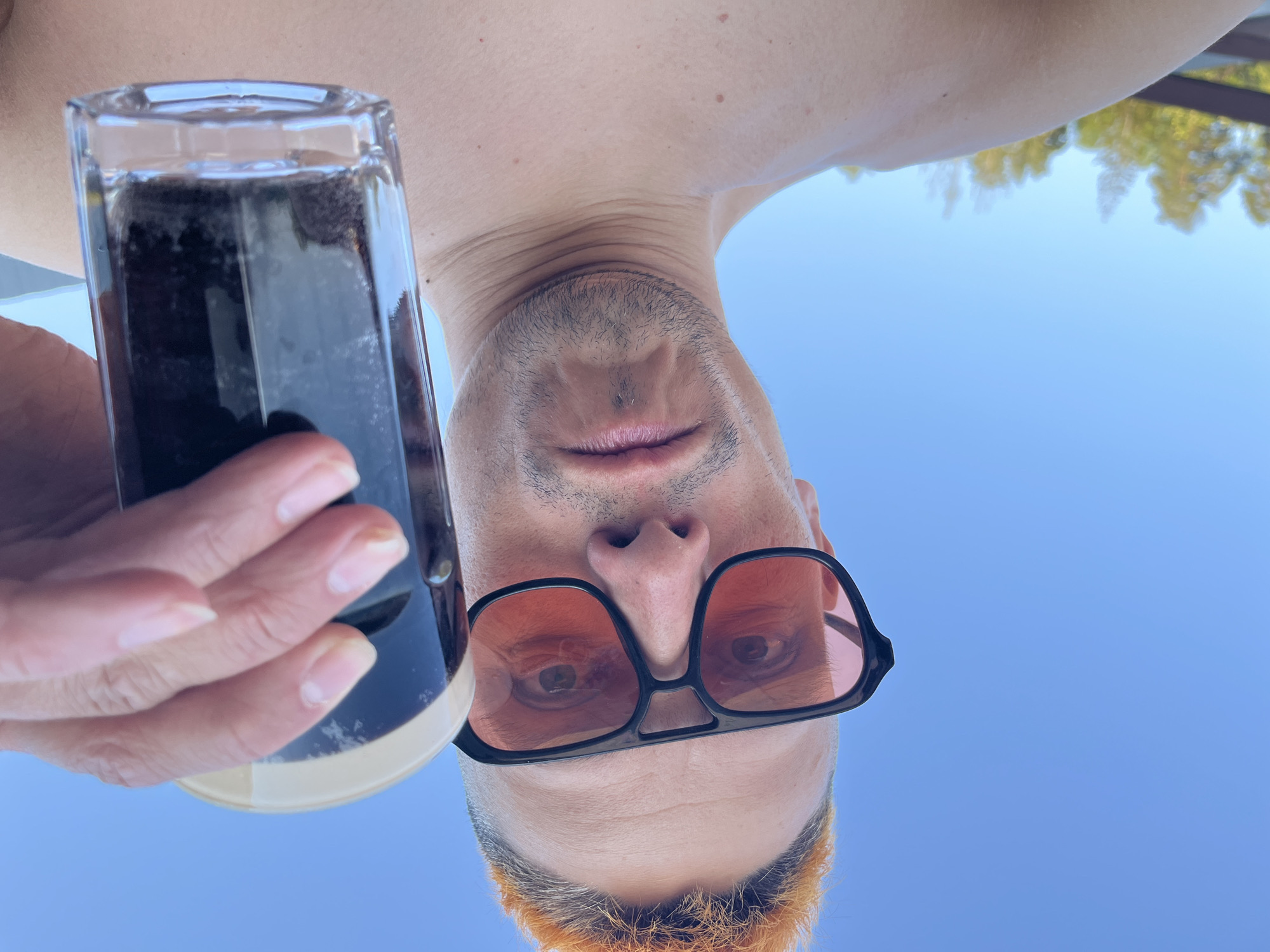 A low-angle shot from Chaikovsky, Russia, shows a man holding a glass of dark liquid, possibly stout or dark beer. The background is clear blue sky. The man has short, dyed orange hair and wears black-rimmed sunglasses with orange lenses. He is fair-skinned. His hand is holding the glass, with visible fingernails. The glass is rectangular and filled with a dark beverage. The camera angle is from below, looking up at the man, obscuring most of his body except for his head, neck, and a small portion of his upper chest.