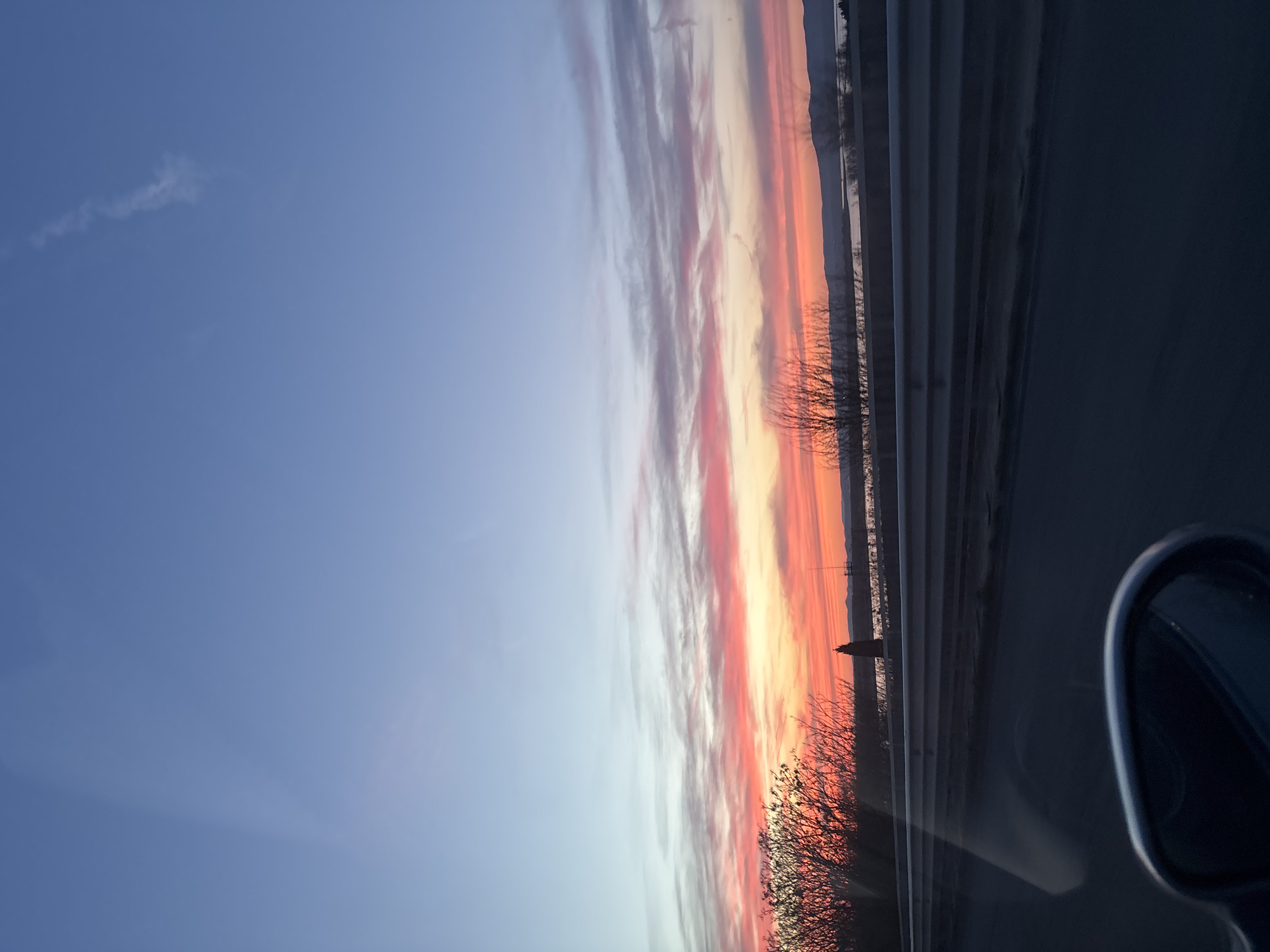 The image shows a sunset with colorful clouds in shades of orange, pink, and white, transitioning to a blue sky on the left side. The photo was taken from inside a vehicle, as a side mirror is visible on the right side of the frame. In the middle ground, there are bare tree branches and roadside barriers. Further in the distance, there appears to be a body of water and a distant coastline. There is a thin spire in the middle of the image. Given the mention of Burgas, Bulgaria, it is plausible that the body of water is part of the Black Sea, and the coastline could be a nearby town or landscape along the coast. The exact location in Burgas cannot be determined from the image alone.