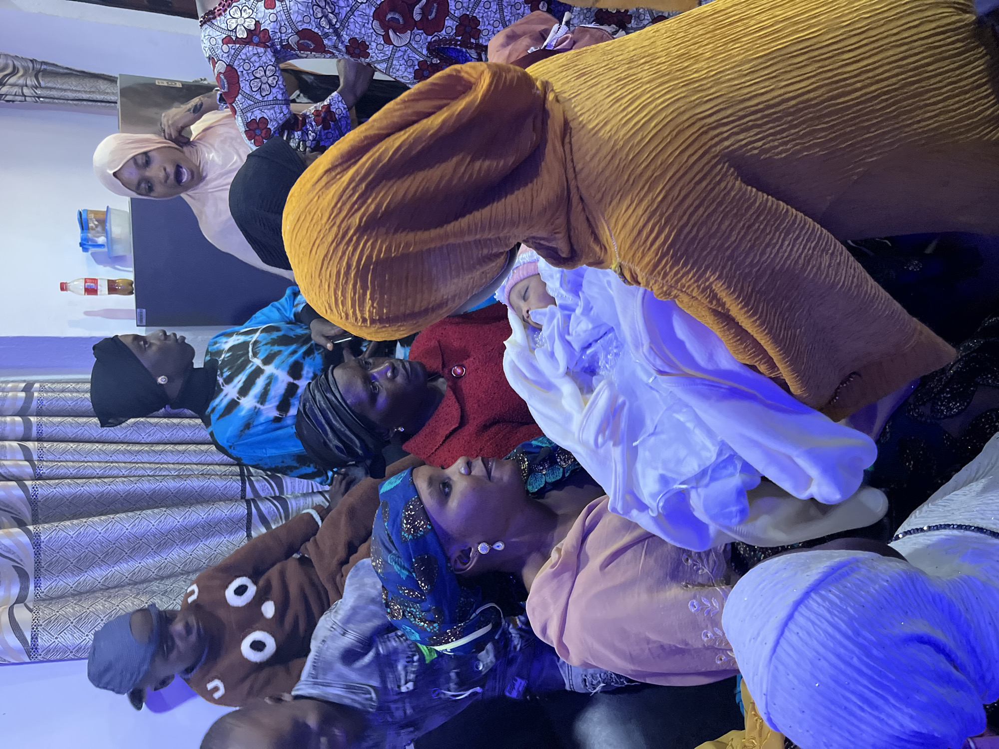 A group of people are gathered indoors, appearing to be celebrating the arrival of a newborn. The scene takes place in a room with patterned curtains, suggesting it is likely a home. A baby is being held by a woman wearing a yellow headscarf, who is facing away from the camera. Other individuals, including women and children, are also present, many wearing headscarves and traditional clothing. The atmosphere appears to be familial and celebratory. The specific location in Jos, Nigeria, cannot be determined from the visual information provided in the image.