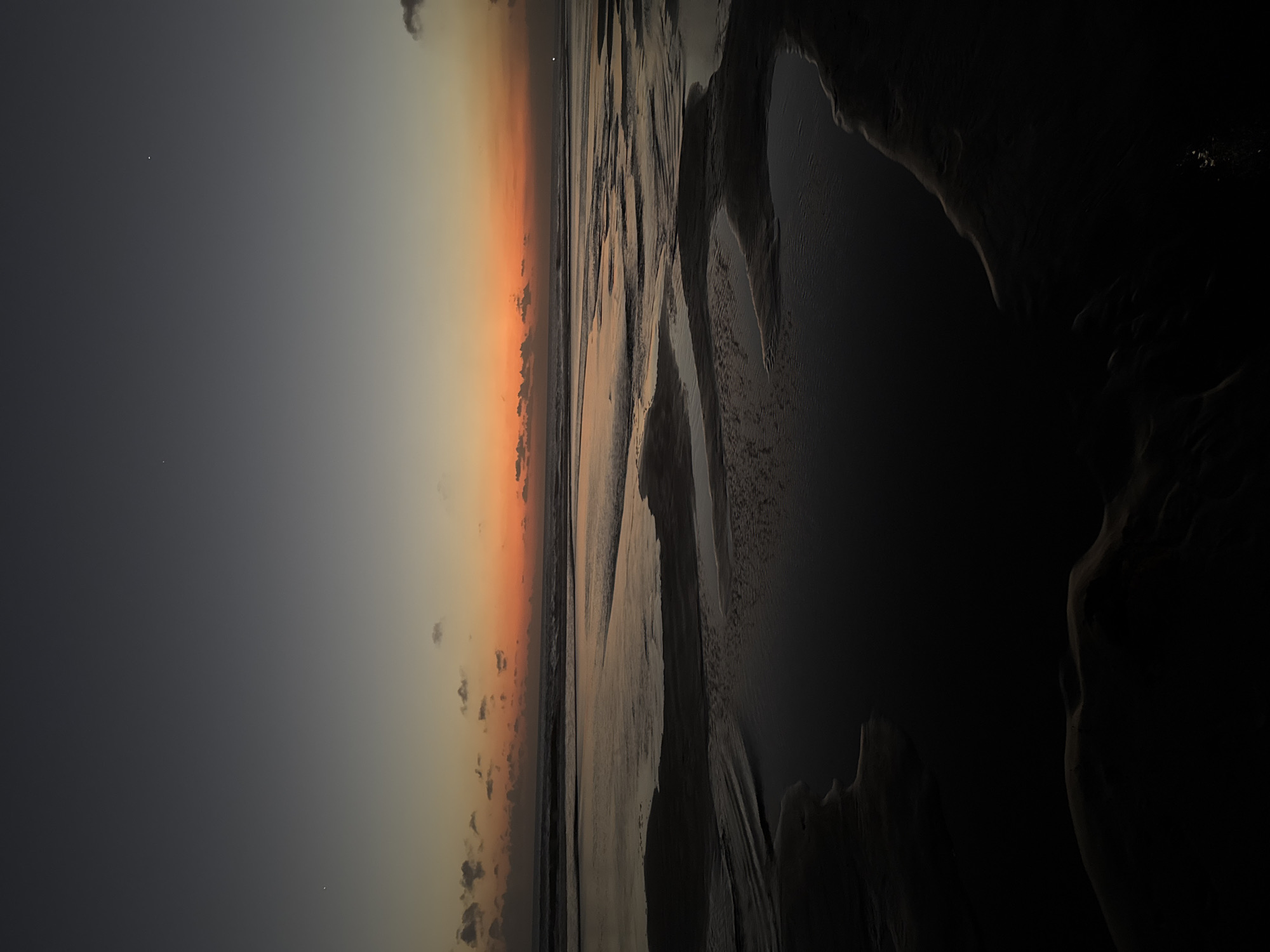 This is a photograph taken at dusk on Kuta Beach in Bali, Indonesia. The scene depicts the wet sand of the beach reflecting the colors of the sunset. The sky transitions from a deep gray at the top to shades of orange and yellow closer to the horizon. Small, dark clouds are scattered across the sky. The ocean is calm, with gentle waves rolling in. The foreground of the image is dominated by dark, wet sand with shallow pools of water, creating a dramatic, low-light effect. There are no people or man-made objects visible in the frame.