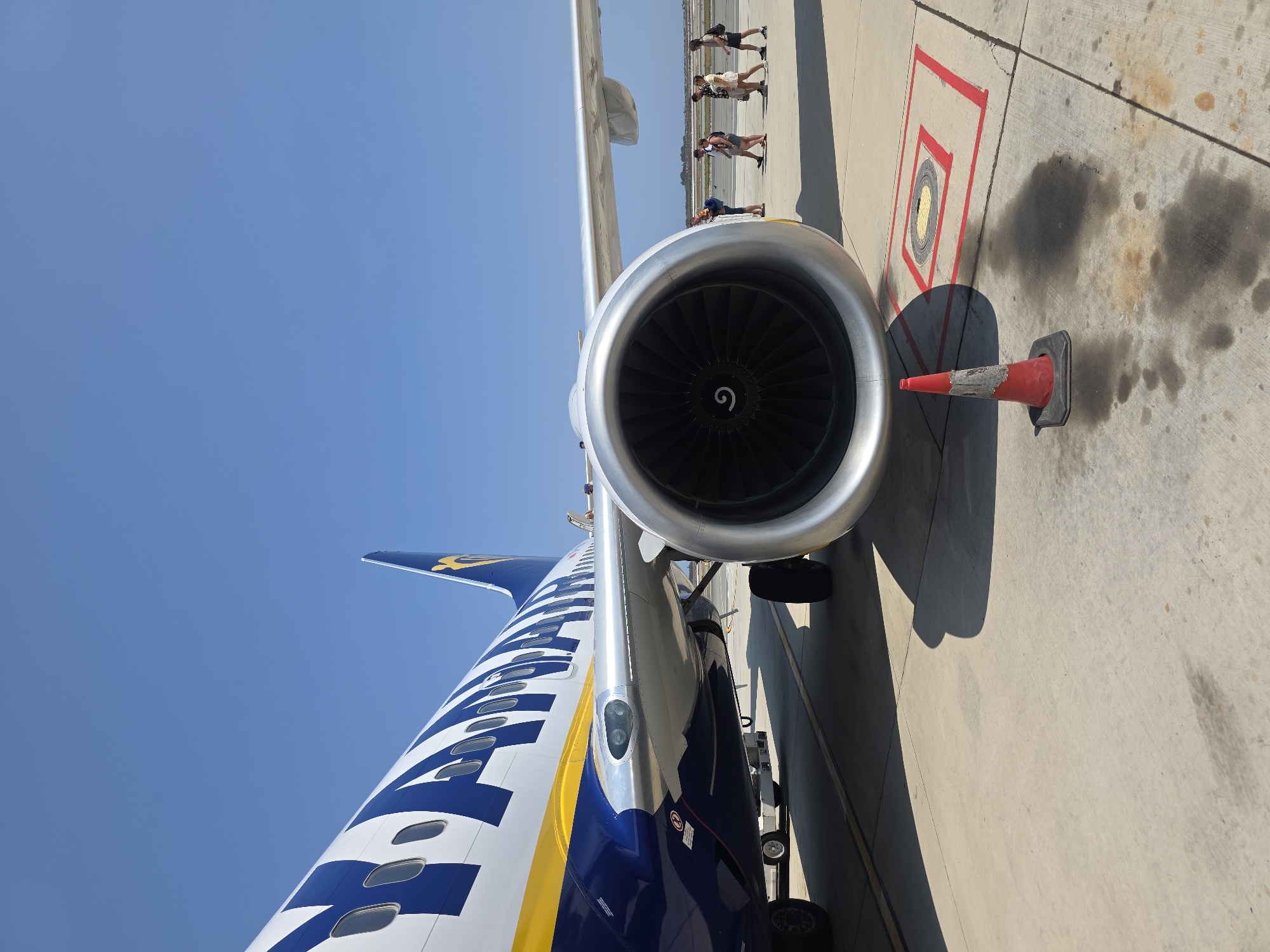 An outdoor, low-angle shot depicts the engine and front fuselage of a parked Ryanair aircraft on the tarmac at Larnaca International Airport in Cyprus. The plane is predominantly white with blue and yellow branding. Its large, silver engine is in the foreground, with its black, slatted interior visible. A red and black traffic cone sits to the right of the engine, casting a shadow. In the middle ground, a group of five people, appearing to be passengers, are walking away from the aircraft towards the right of the frame. They are dressed in casual summer clothing. The background shows a clear, bright blue sky and a distant treeline. The tarmac is concrete, with painted yellow and red lines marking an area near the traffic cone. The scene is well-lit by natural sunlight.