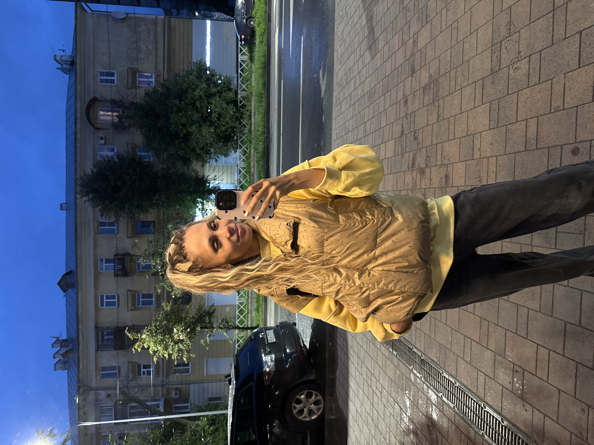 A young woman with blonde hair in a ponytail is taking a selfie with her phone. She is wearing a yellow sweatshirt and a tan puffer vest, and dark gray jeans. She is standing on a wet, paved sidewalk with a metal drain grate. Behind her is a street with a black SUV and a multi-story building with yellow walls. The sky is dark blue, indicating it is either early morning or evening.