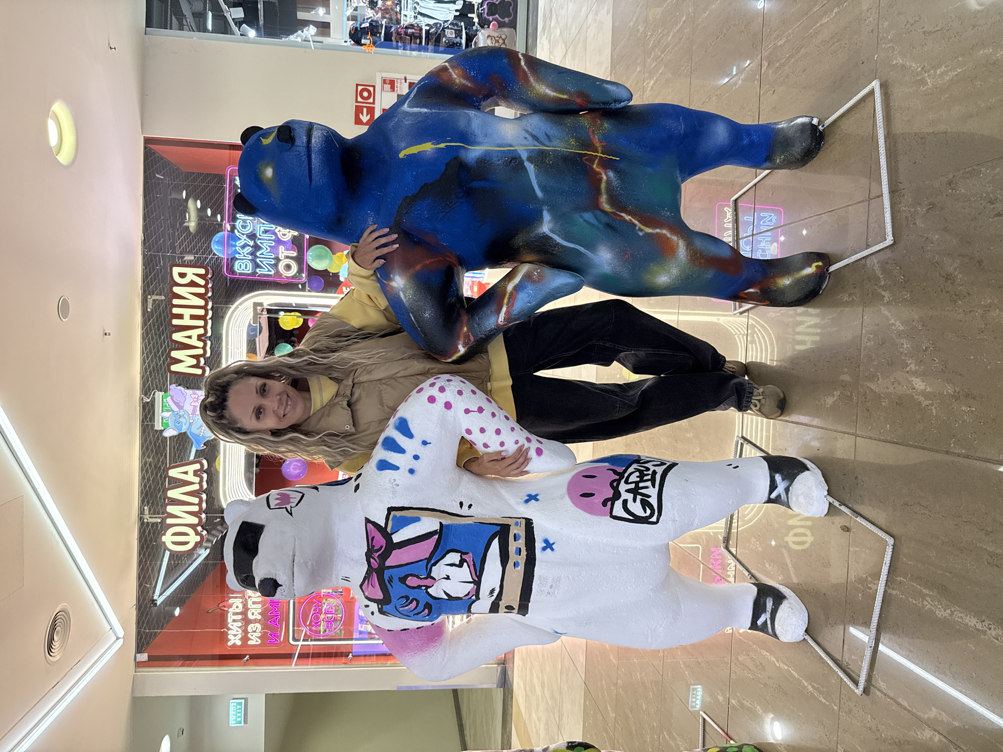 In Yaroslavl, Russia, a woman stands between two large, stylized bear sculptures in a brightly lit indoor shopping mall. The woman, with blonde, wavy hair pulled back, is wearing a tan puffer vest over a yellow sweatshirt and dark jeans. She smiles directly at the camera, with her left arm resting on the white bear's shoulder and her right hand on the blue bear's shoulder.
The white bear sculpture is painted white and adorned with colorful artwork, including a pink bow and a depiction of a person in a framed picture on its back, along with pink dots and blue X's. It wears black and white sneakers. The blue bear sculpture is primarily blue with abstract, colorful spray-painted accents in red, yellow, and green, giving it a cosmic or marbled appearance. It also wears black boots.
Behind the sculptures, neon signs are visible in Russian. One sign reads "ФИЛА МАНИЯ" (Phila Mania) in large, yellow letters, and another reads "ВКУСНО ИМПОРТ ОТ" (Tasty Import From) in smaller letters with a blue outline. Various balloons are also visible in the background, suggesting a festive or promotional event. The floor is tiled in a light brown color, and metal frames are placed around the base of each bear sculpture. The overall environment suggests a retail space or a public area within a shopping center.