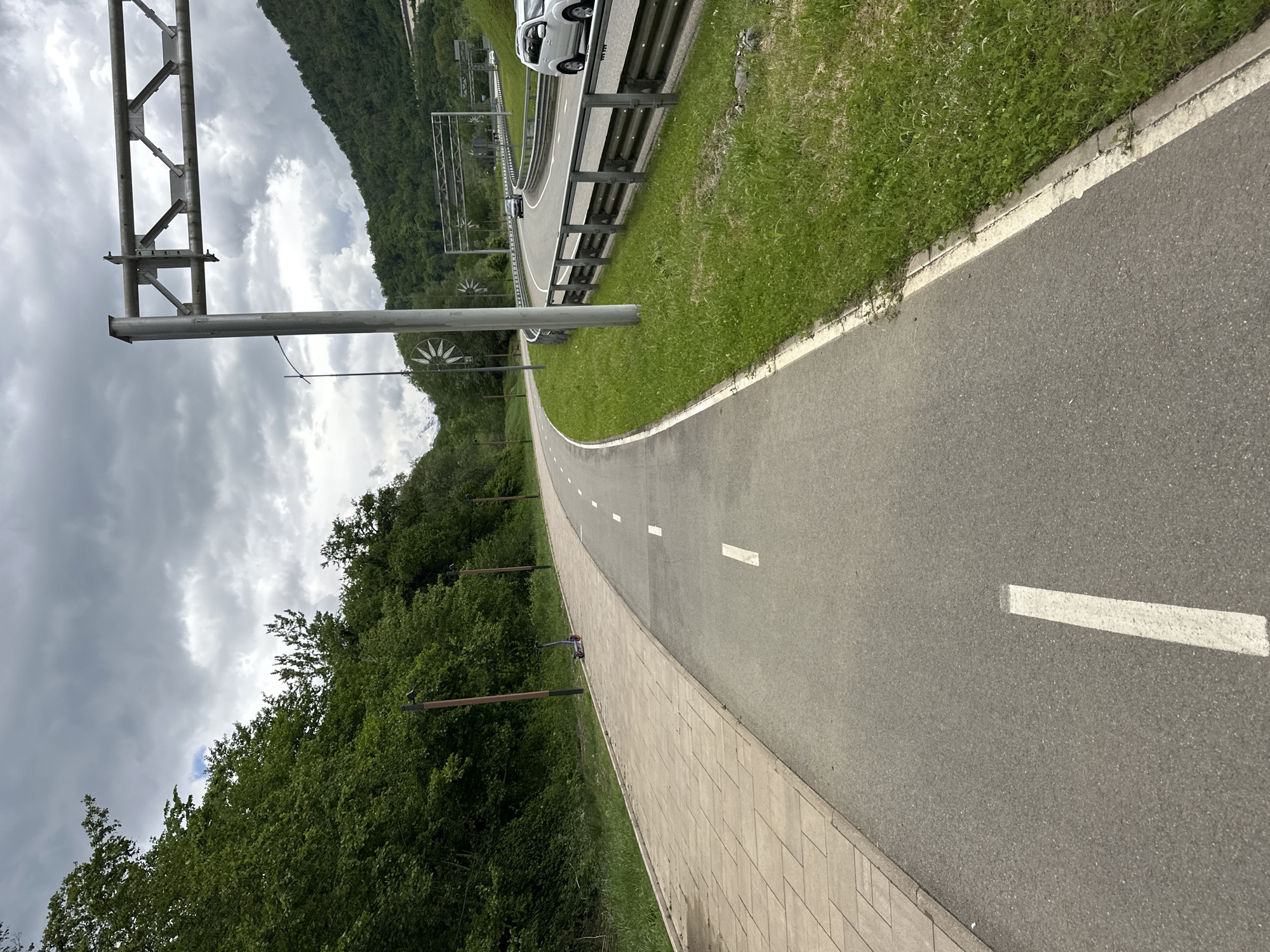 The image depicts a paved pathway in Krasnaya Polyana, Russia, alongside a road with a metal guardrail and occasional passing vehicles. The pathway is marked with white dashed lines. Dense green vegetation covers the adjacent hillside. Overhead are metal structures, possibly for power lines or cable cars. The sky is overcast with dark clouds. A paved brick border separates the pathway from the bordering vegetation. Tall trees border the other side of the pathway.