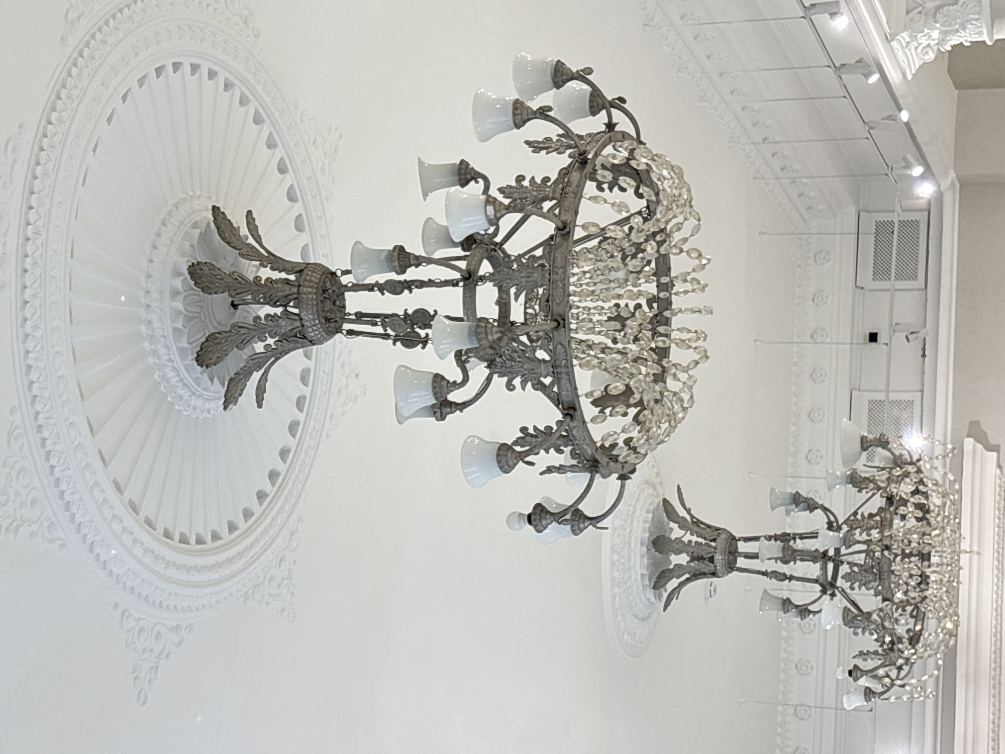 The photograph captures the interior of a space with a focus on decorative lighting and architectural details. Two ornate chandeliers, featuring multiple frosted glass lamp fixtures and strands of crystals, hang from the ceiling. The ceiling is adorned with a circular, layered plaster medallion surrounding each chandelier base. The background reveals additional architectural elements, including ceiling panels, moldings, and a visible air vent. The photograph might have been taken in a high-ceilinged room with intricate detailing, potentially a historic building in Perm, Russia.