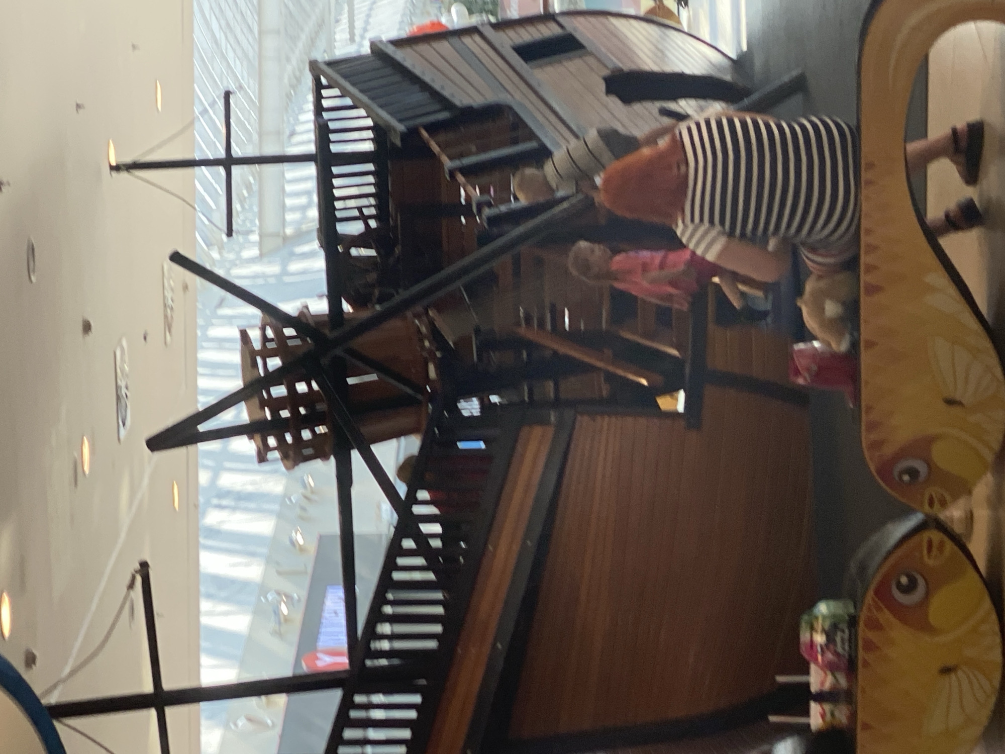 The image shows a large, wooden, pirate-ship-themed playground structure inside a building with a high, glass-covered ceiling. Several people, including children and an adult woman with red hair, are present on and around the ship. One child in a pink shirt is on a staircase, and another child in a striped shirt is climbing the stairs. The adult woman, wearing a striped shirt and skirt, is sitting on a bench in the foreground. The bench has a decorative design of two yellow fish. In the background, the interior of a building is visible, including shops and bright lighting from the ceiling. The location is identified as Novokuznetsk, Russia.