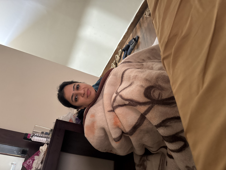 A person with dark hair and eyes is lying in bed under a patterned blanket. The blanket has large, swirly, dark brown designs. Part of a dark wood nightstand is visible with a partially obscured water bottle and what appears to be tissue. In the background, there are cream-colored walls and ceiling. A small rack with sandals and shoes are mounted on the wall. A golden curtain is partially visible.