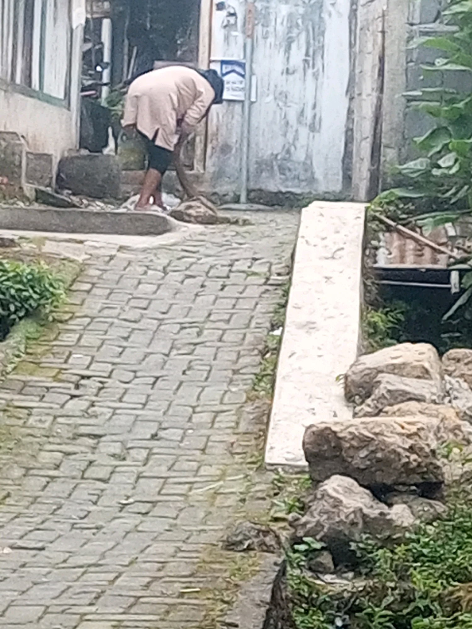 In Andongsili, Indonesia, a person wearing a pale pink hooded garment and dark pants is bent over, possibly cleaning or gathering something from the ground. The setting appears to be a pathway paved with interlocking bricks, sloping downwards. To the right, a concrete ramp or step leads up, bordered by rocks and lush green vegetation. The background shows a weathered wall and a building.