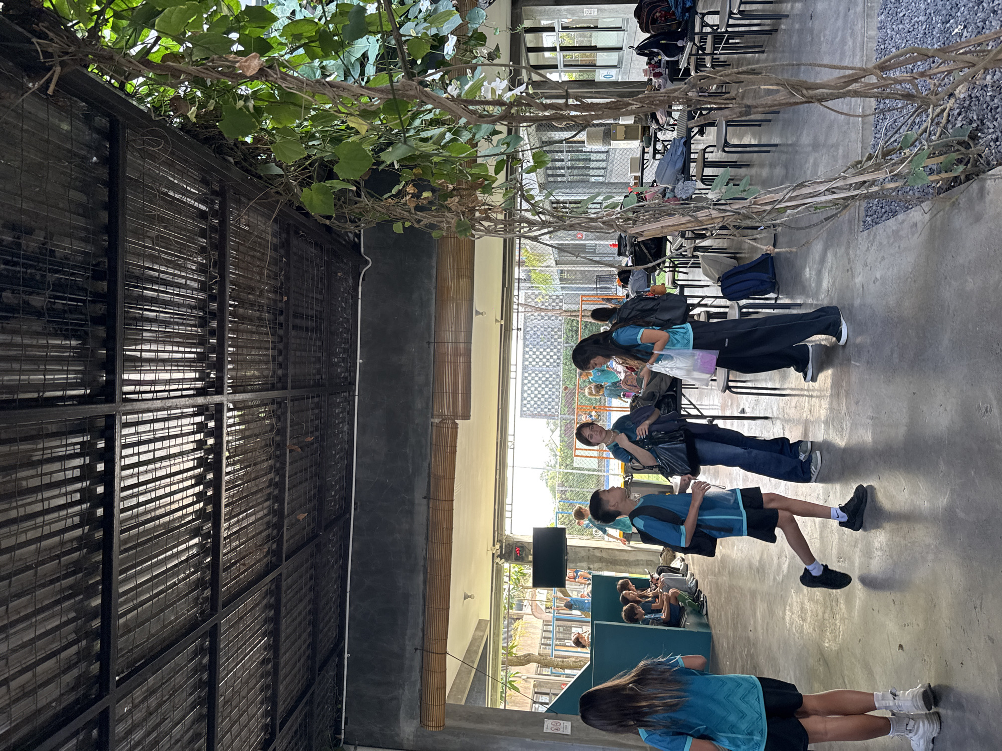 The image depicts an outdoor area, possibly a school or community center, in North Kuta, Indonesia. The foreground shows several individuals, including two young girls in turquoise tops and black shorts, one walking away from the camera and the other facing it. A boy in a turquoise t-shirt and black shorts walks to the right, while two women stand in the center talking, one in a black jacket and the other in a turquoise top and black pants. In the background, more people are seen sitting at tables and standing. The environment features a covered seating area with a metal roof, concrete floors, and a prominent vine-covered structure. The general atmosphere suggests a casual gathering or a break time.