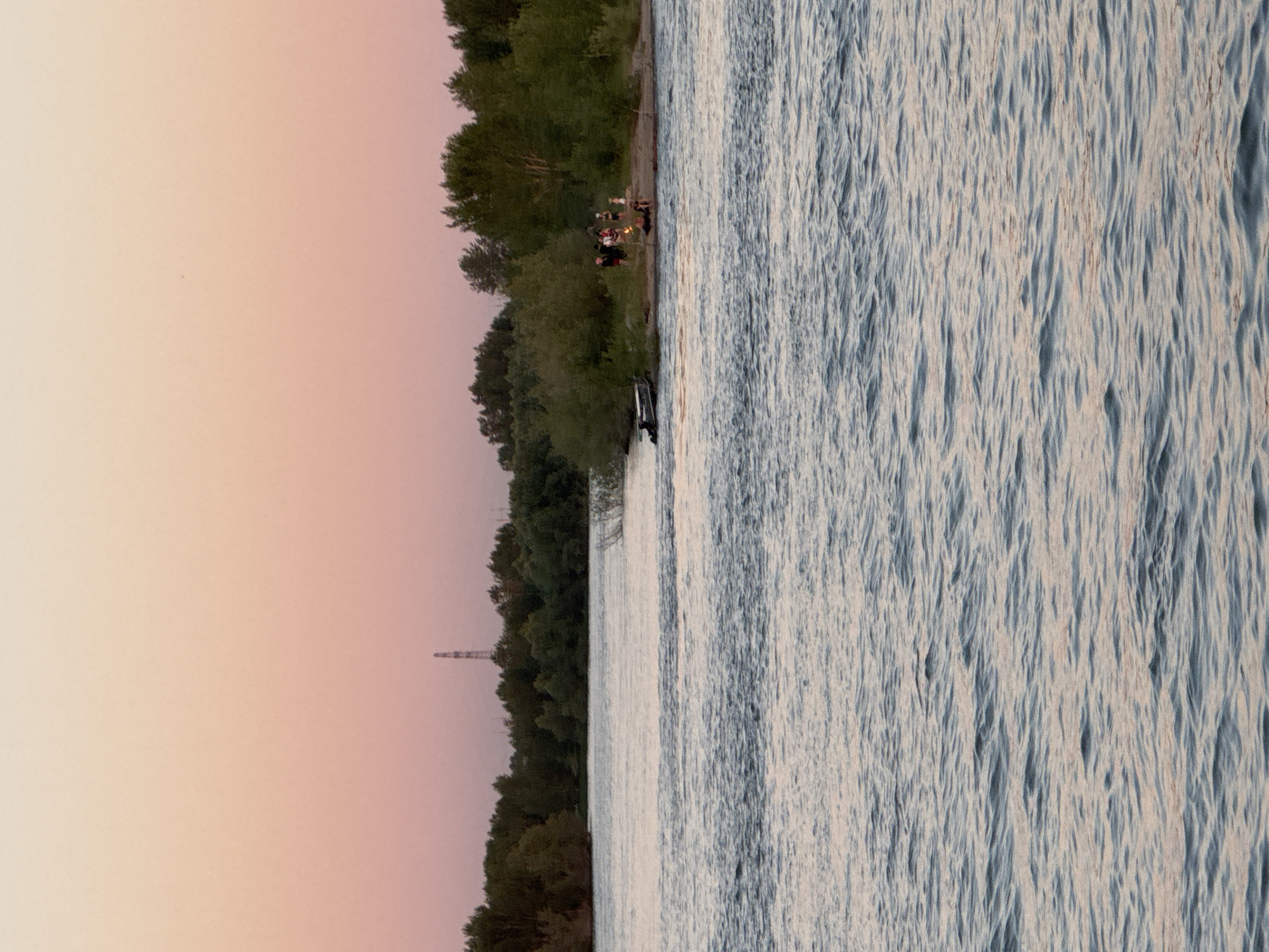 The image depicts a serene evening scene by a body of water, likely a river or lake, with a gentle breeze creating rippling textures on the water's surface. The water occupies the right half of the frame, reflecting the soft hues of the sky. The left side of the image transitions from a pale pink sky, indicative of sunset or sunrise, to a dense line of dark green trees that form the shoreline. A distant industrial structure, possibly a tower or pylon, is visible on the far left, silhouetted against the sky.
On the grassy bank nestled amongst the trees, a small group of people are gathered around a campfire. Their figures are small and somewhat indistinct, but they appear to be enjoying each other's company, with some seated and others standing. The campfire itself is a bright point of light, suggesting warmth and conviviality. Near the water's edge, a dark object, possibly a vehicle or a boat, is partially submerged or parked close to the shore. The overall atmosphere is peaceful and relaxed, hinting at a casual gathering or a moment of respite. The time of day is clearly evening, given the soft, warm light and the presence of the campfire. The location is indicated to be Novosibirsk, Russia, which conjures images of similar natural landscapes near the city. No visible text is present in the image.