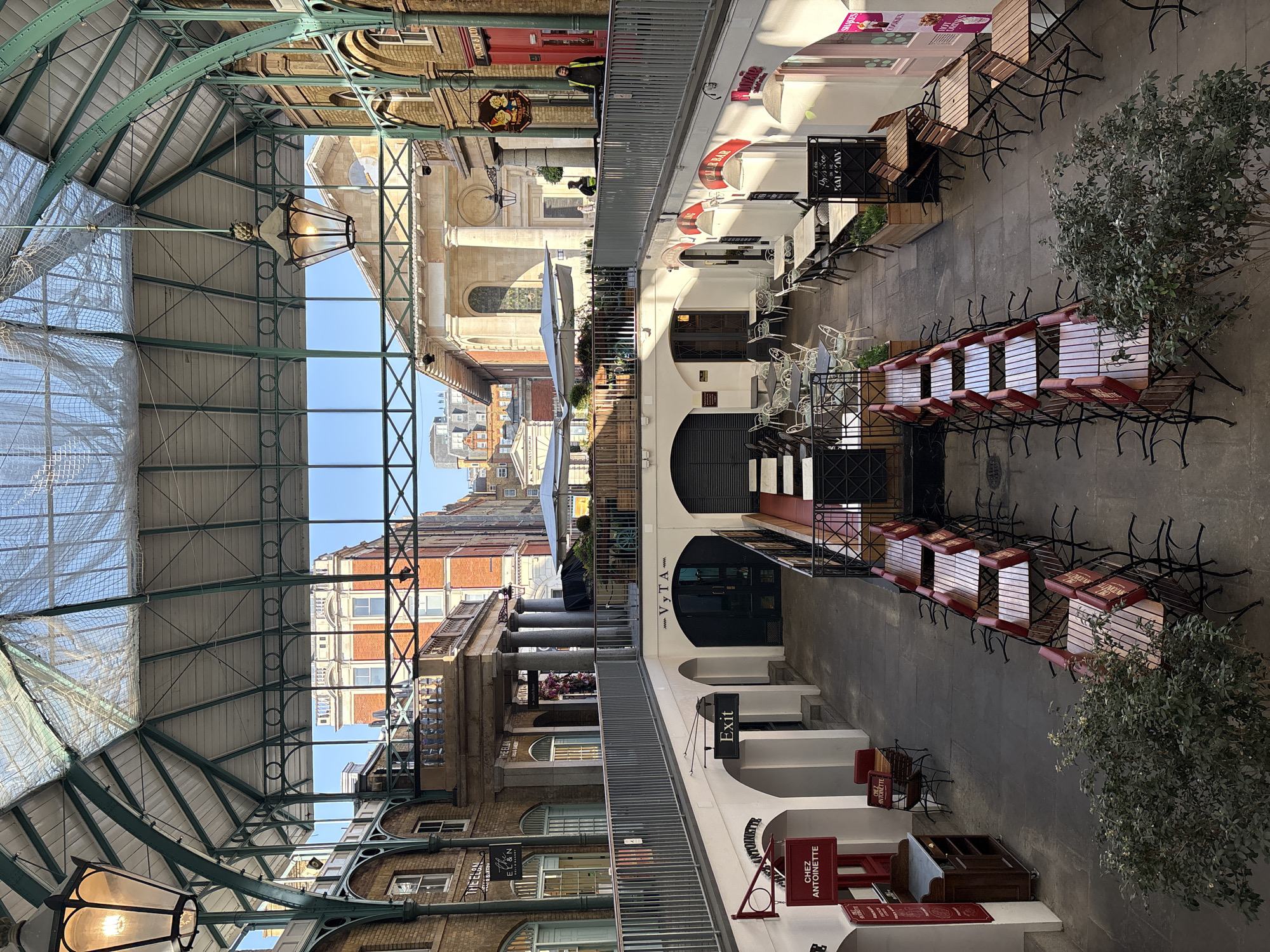 The image depicts a grand, covered market hall or arcade, likely in a European city given the architectural style. The perspective looks down the length of the hall, revealing a high, arched glass and green metal framework roof through which a clear blue sky is visible. Bright sunlight streams through the roof, creating strong shadows and illuminating the scene.
On the ground level, numerous outdoor dining areas are set up on a paved surface, featuring a mix of white metal chairs and dark metal chairs with wooden or red slats. Many chairs are stacked or neatly arranged, suggesting the area might be empty, possibly early morning, off-peak hours, or closed. Large decorative hanging lanterns are spaced along the ceiling.
Along both sides of the hall are multi-story buildings housing various establishments. These buildings display classical architectural elements such as columns, arched doorways, and brick facades. Planters with green foliage are strategically placed, adding natural touches to the urban setting.
Very few people are present. In the distance, two individuals, one wearing a high-visibility vest, can be faintly seen near a shop entrance, possibly staff or maintenance. No significant activity or event is taking place, contributing to the tranquil atmosphere.
Visible text on signs includes: "EXIT," "CHEZ ANTOINETTE," "THE ELAN" (or "EL&N"), "THE BALCONY," "BAR," "VYTA," and a partial "CHURCH & CO." Another sign mentions "TAKE AWAY," and banners show "SWEETS" and "SHAKES." The scene suggests a refined commercial or leisure area.