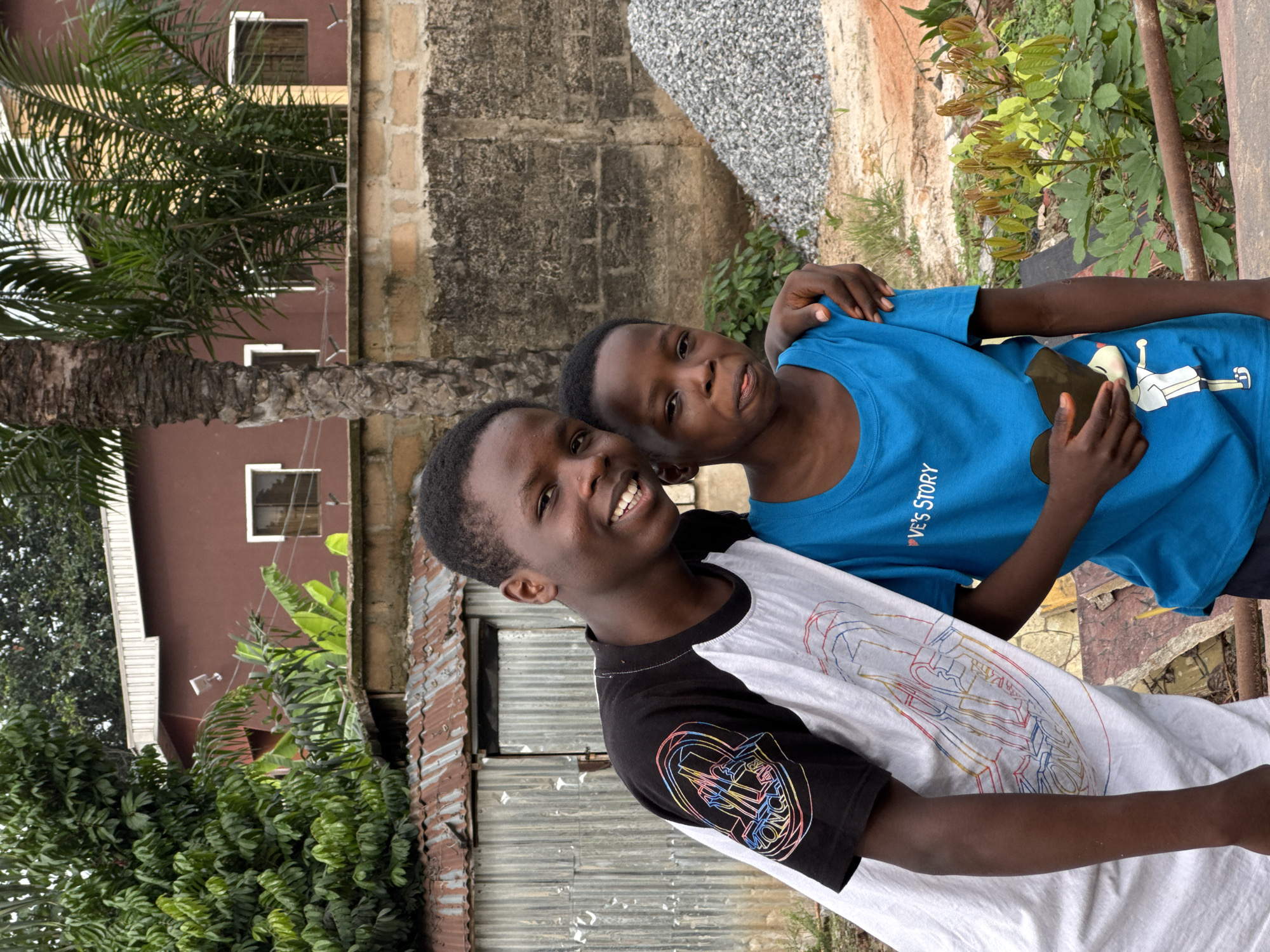 Two African children are standing outdoors in Orifite, Nigeria. The taller child wears a black and white shirt and smiles broadly, with a smaller child in a blue shirt wrapping an arm around his shoulder. Behind them is a mix of vegetation, including a palm tree, a corrugated metal fence, and concrete buildings. A pile of gravel or rocks is also visible.