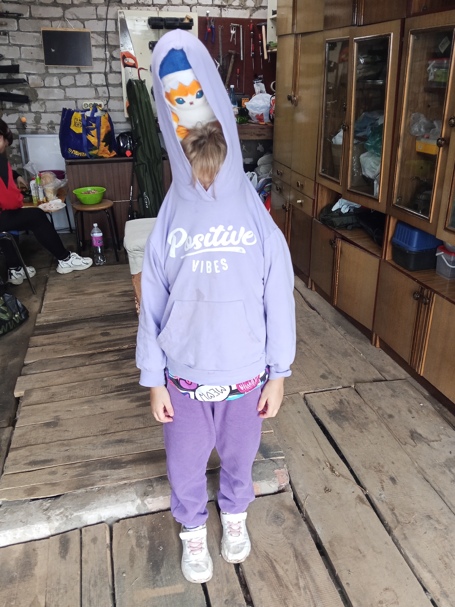 A person stands in what appears to be a garage or workshop with wooden floors and brick walls. The person is wearing a purple hoodie, purple sweatpants, and white sneakers. The hoodie is pulled up over their head, and a stuffed animal with orange and white fur and blue accents is visible peeking out from the hood. The stuffed animal resembles a cat with large eyes and pointy ears. The hoodie has the words "POSITIVE VIBES" printed on the front in white lettering. The person is standing on a wooden floor, and in the background, there is a large wooden cabinet with glass doors, a tool rack on the wall, and several bags and containers. The overall scene suggests an indoor, cluttered environment.