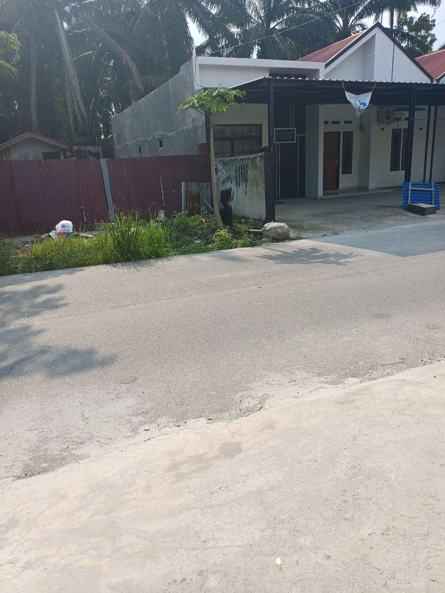 The image depicts a residential street in Dumai City, Indonesia, under bright daylight. The scene features a white house with a dark-colored roof and a covered parking area. Adjacent to the house is a corrugated metal fence, partially covered by overgrown grass and vegetation. In front of the fence, a white plastic bag and some debris are visible. The street itself is paved and appears to be in good condition, with shadows of trees cast upon it. In the background, palm trees and other tropical foliage are abundant, characteristic of the region. A blue plastic structure, possibly for seating or storage, is placed near the edge of the property. The overall impression is that of a typical suburban or residential area in Indonesia.