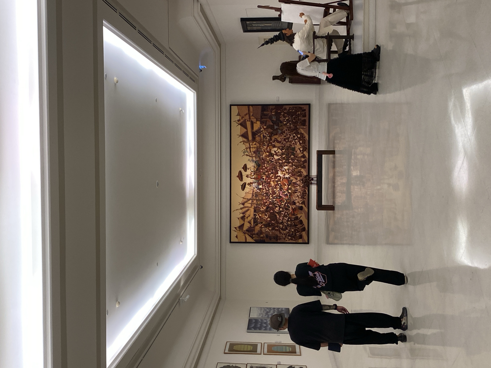 A gallery space in Bangkok, Thailand features a large, vertically oriented historical painting with a brown palette depicting a crowded scene, prominently displayed on a white wall. In the foreground, two individuals are viewing artworks; the first person, closer to the painting, is a woman wearing black, while the second, a man, also in black attire, stands slightly behind. To the upper right of the large painting, there is a seated figure positioned near an easel, possibly part of a display. The gallery space is well-lit with recessed lighting along the ceiling, which is a stark white, as are the walls. A row of smaller framed images is visible below the main painting. The flooring is a polished, light-colored surface, reflecting the light.