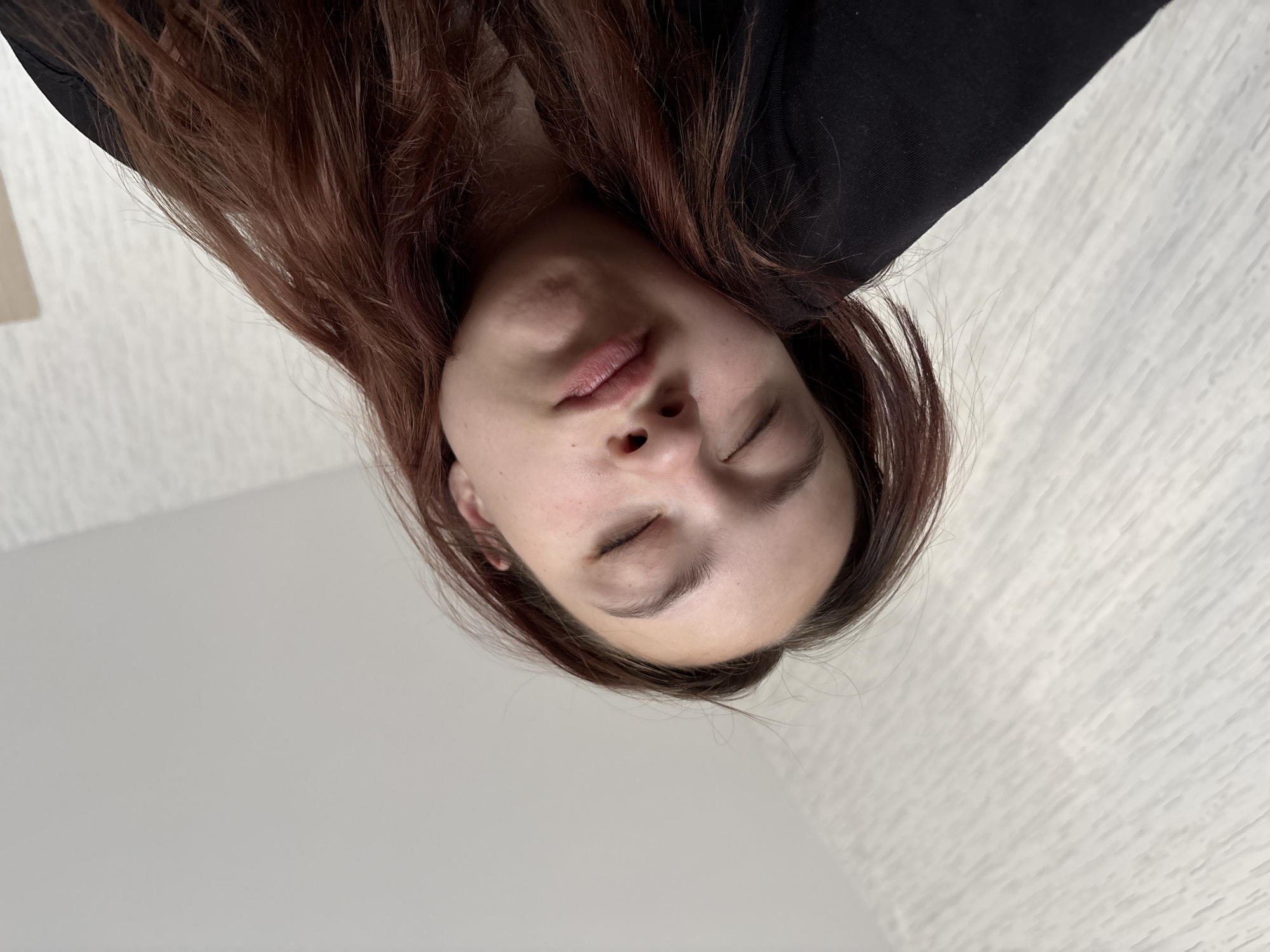 A close-up, inverted image shows a woman with closed eyes. She has long, reddish-brown hair. She is wearing a black top. The background is a neutral, slightly textured surface. No specific location information about Perm, Russia, is discernible from the image.