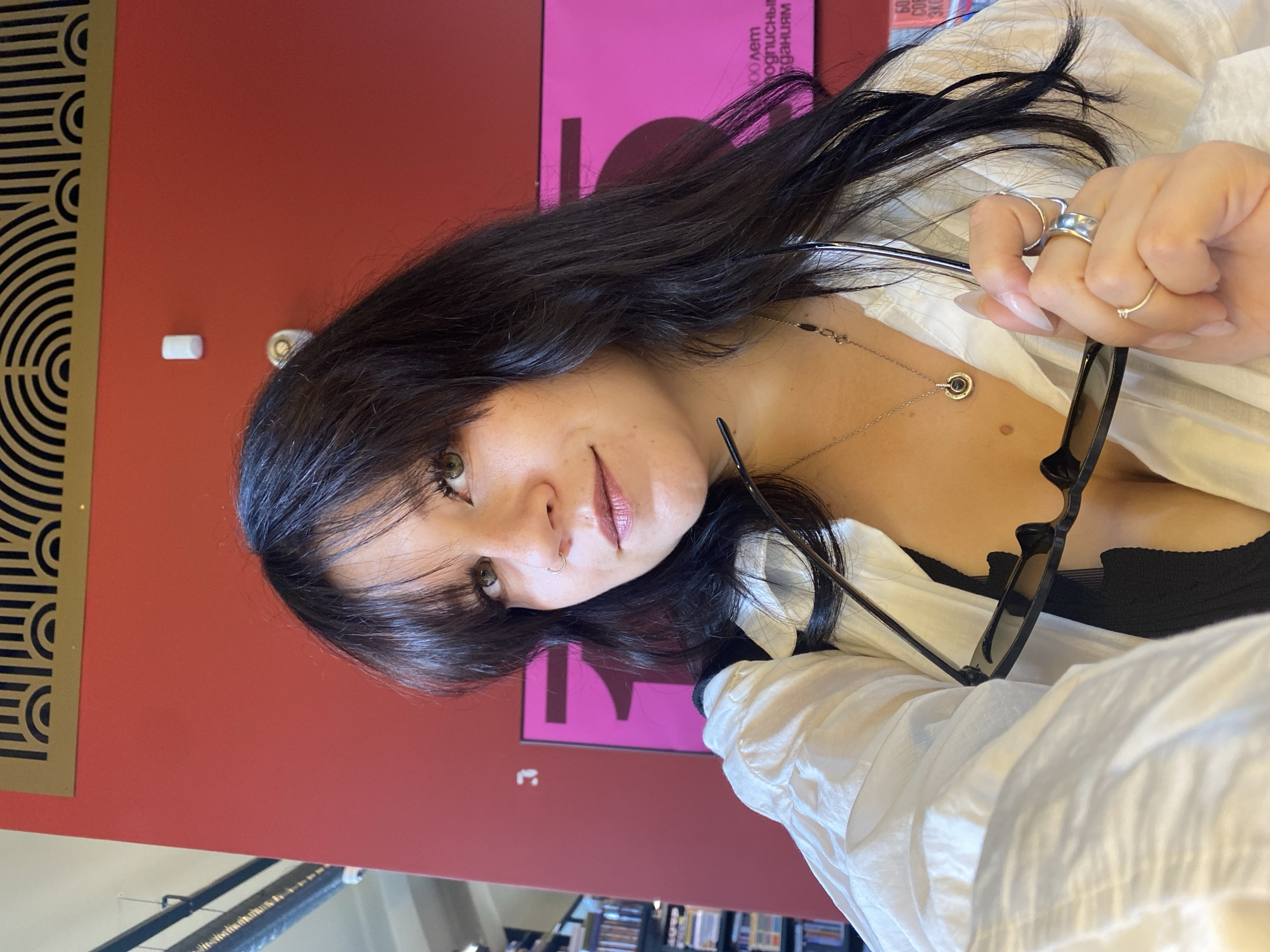 A young woman with long, dark hair and bangs is taking a selfie indoors, holding a pair of sunglasses. She is wearing a white button-down shirt, partially unbuttoned, revealing a black top underneath. She has a silver necklace with a pendant and several rings on her fingers. The background features a vibrant red wall with a decorative gold and black art deco-style element at the top, and a large pink sign with text and a stylized number "5" on the right. The overall setting suggests an interior space, possibly a museum, library, or gallery, given the visible bookshelves and signage which could indicate a cultural institution in Saint Petersburg, Russia, given the Cyrillic text on the pink sign.