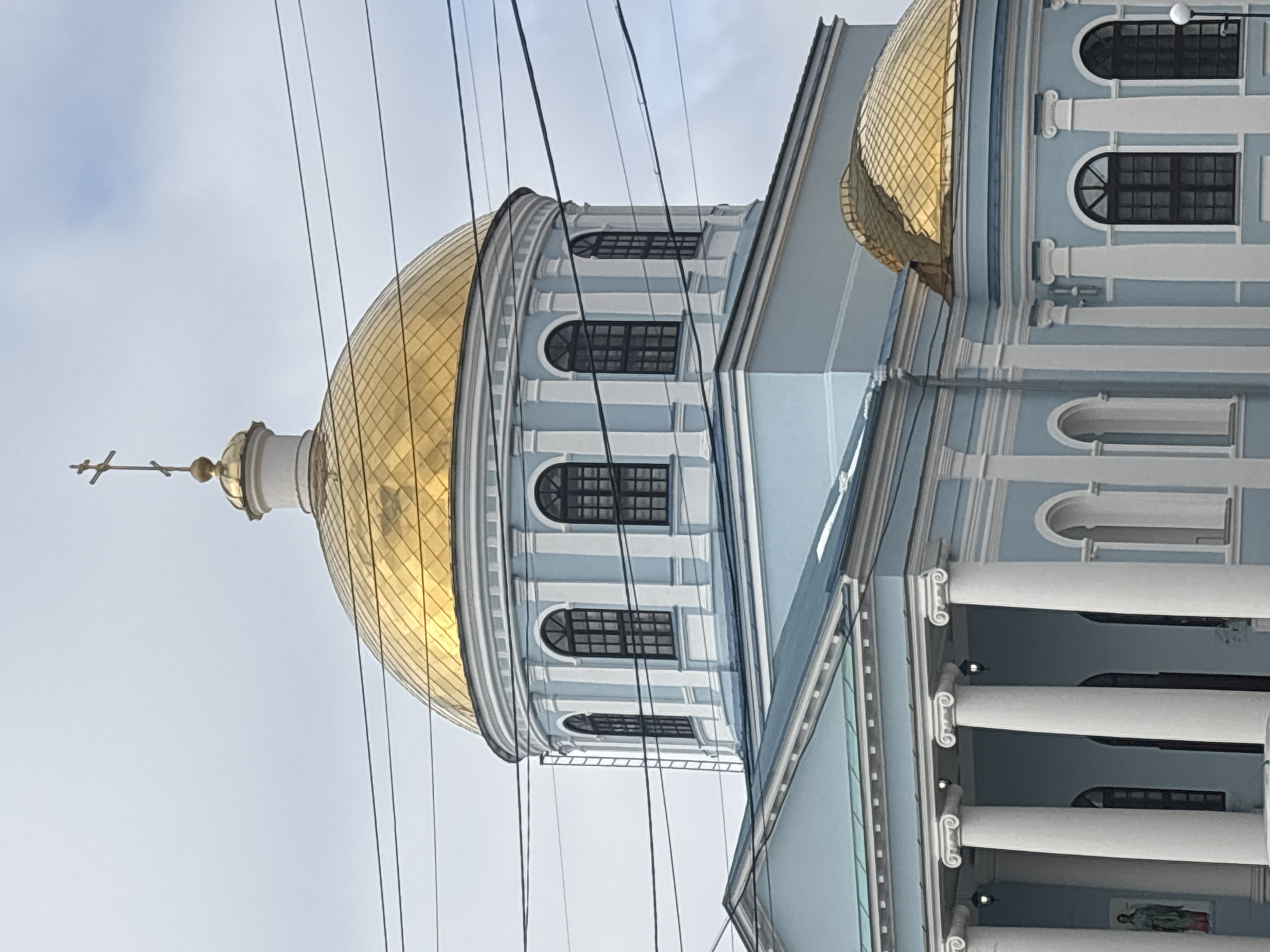 The image shows a close-up view of the architectural details of the Blagoveshchensky Cathedral, located in Votkinsk, Russia. Predominantly light blue and white, the building features a large, golden dome topped with a cross. Windows are visible on the dome structure. Power lines are visible in the foreground, cutting across the view of the architecture. Columns are visible on the cathedral. The sky is overcast.