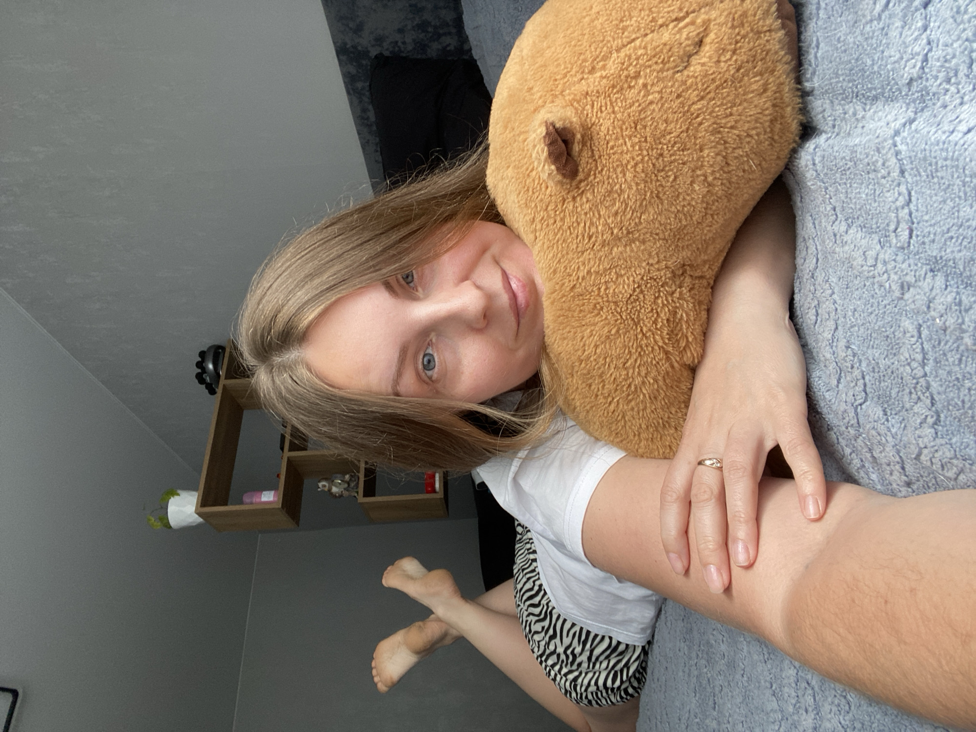 A woman with fair skin, blonde hair, and blue eyes is lying on a light blue, textured surface, likely a bed, hugging a brown stuffed animal. She wears a white t-shirt and zebra-striped shorts. A gold ring is visible on her left ring finger. In the background, there is a gray wall, a wooden shelf with small decorative items and a potted plant. The location is not discernible as Noviy, Russia based on the image content.