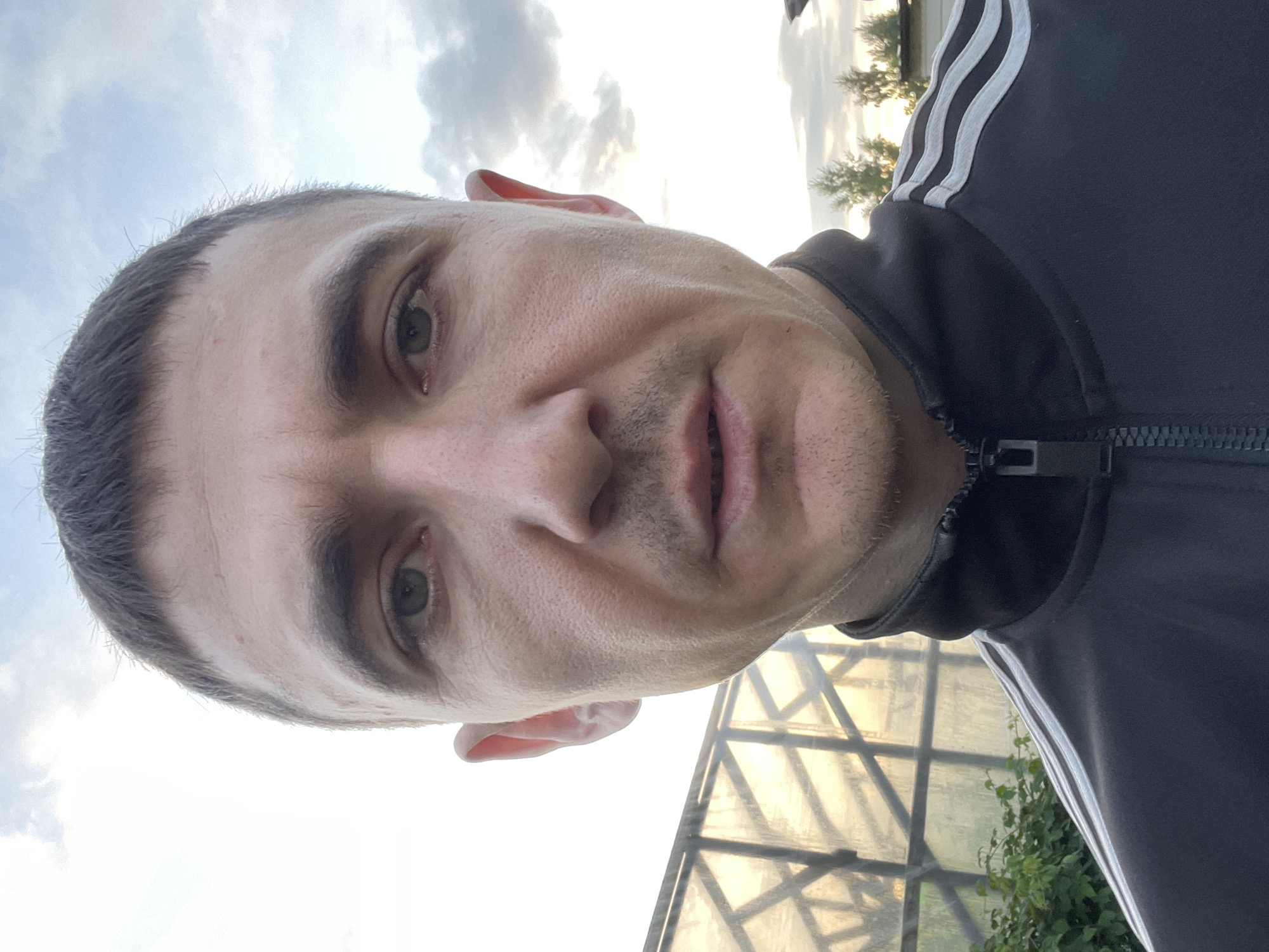 A man with short dark hair and green eyes, wearing a black tracksuit with white stripes on the shoulders, is captured in a close-up selfie. The man's face is centrally positioned, with his gaze directed towards the camera. Behind him, a greenhouse with a reflective surface and a structure of intersecting bars is visible. The sky above is partly cloudy with soft light. The environment suggests an outdoor setting, possibly a backyard or a rural area. The specific location within Laishevo, Russia, is not discernible from the visual elements in the image.