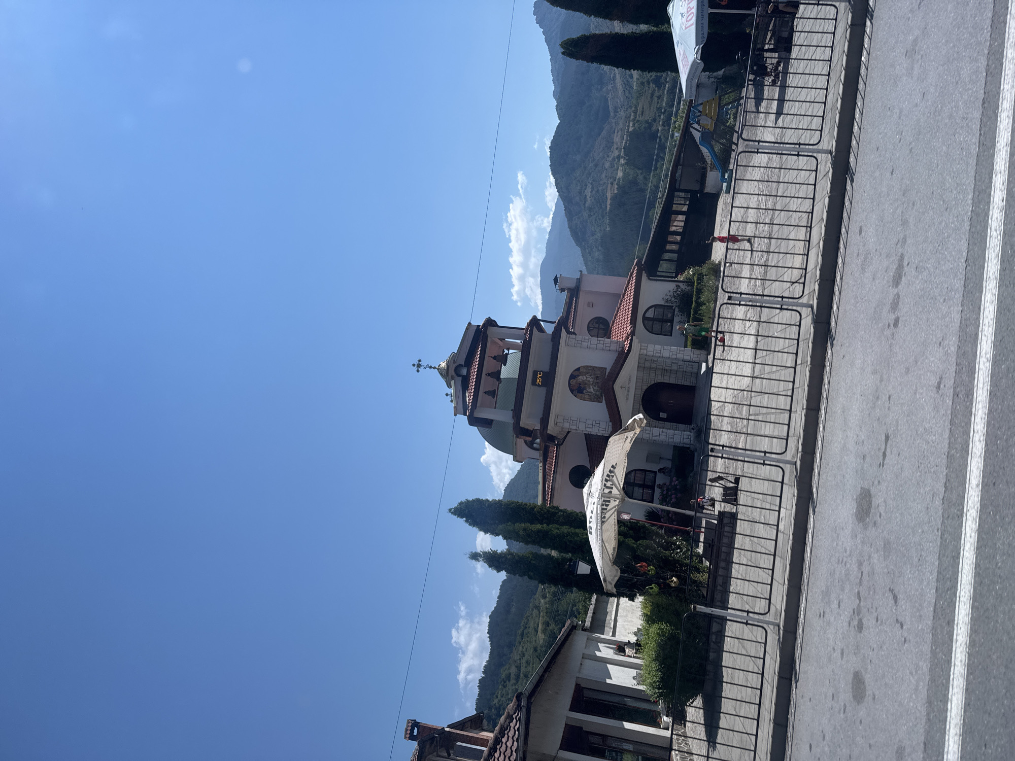 This is a ground-level, eye-level, outdoor, daylight, full-shot photograph of a church in Mesta, Bulgaria. The church is a prominent pink and white structure with a green domed roof, topped with a cross. It has multiple levels, arched windows, and decorative architectural elements. A digital thermometer on the church facade displays "25°C". In front of the church, there is a paved courtyard enclosed by a black metal fence. An open white umbrella with visible text is positioned in the courtyard, and several cypress trees are planted nearby. To the left of the church, a smaller white building with a red tile roof is visible. In the background, a playground structure with a slide and swings can be seen. Further in the distance, lush green mountains with scattered trees form the backdrop under a clear blue sky with a few wispy clouds. A paved road with white lane markings is in the foreground, and a sidewalk with a curb separates the road from the fenced area. There are at least three individuals visible: one person in a red shirt standing in the courtyard, another person in green shorts to the left, and a third person, possibly a child, in the distance near the playground. The overall scene depicts a sunny day in a small town.