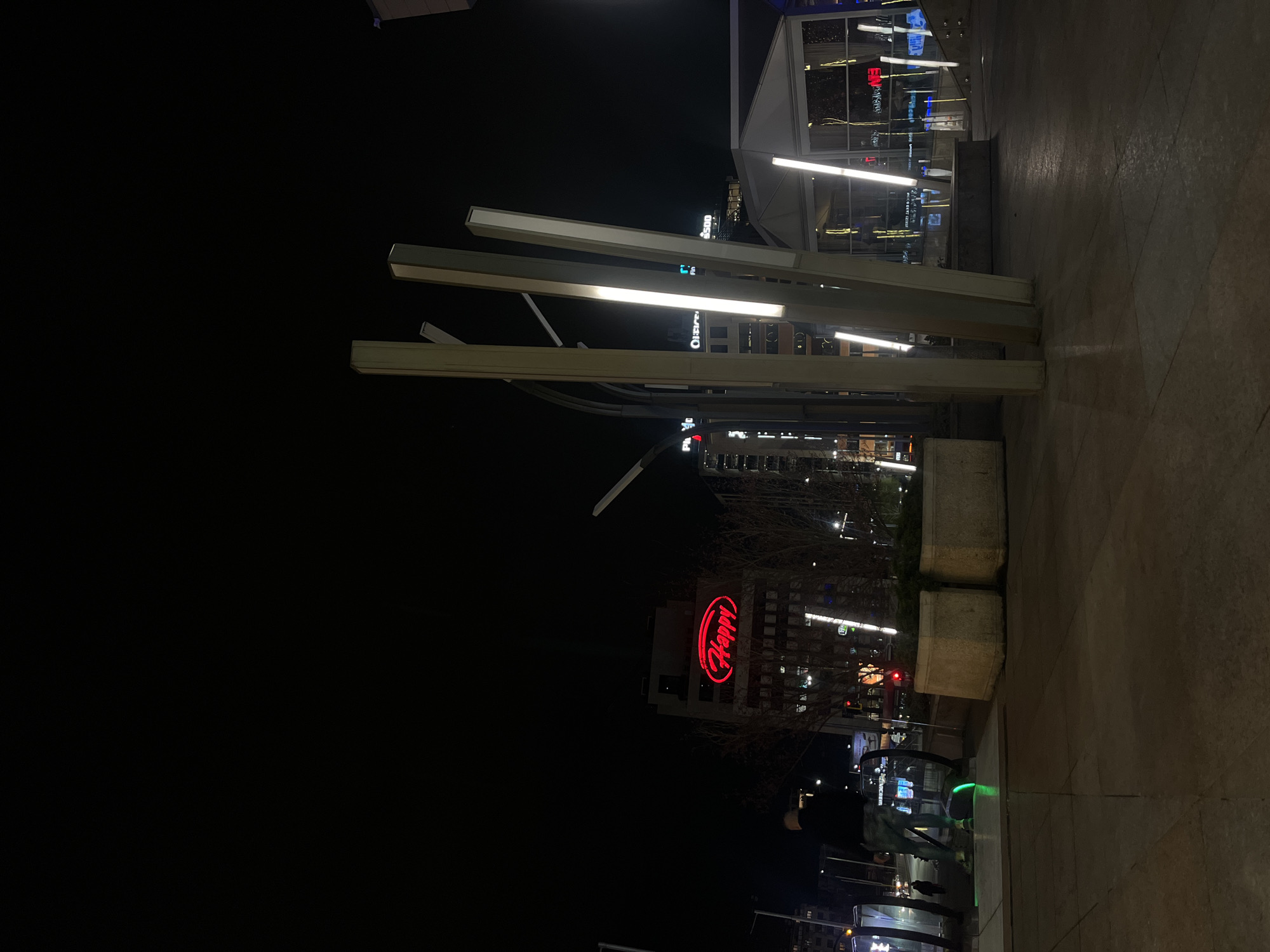 The image depicts a night scene in Sofia, Bulgaria. In the foreground are modern light fixtures constructed of parallel, illuminated bars, and planter boxes. In the midground, the "Happy" restaurant sign glows red on a building, surrounded by other structures and lights. In the background, the skyline of Sofia is visible. The scene is generally dark with light concentrated around the illuminated elements.