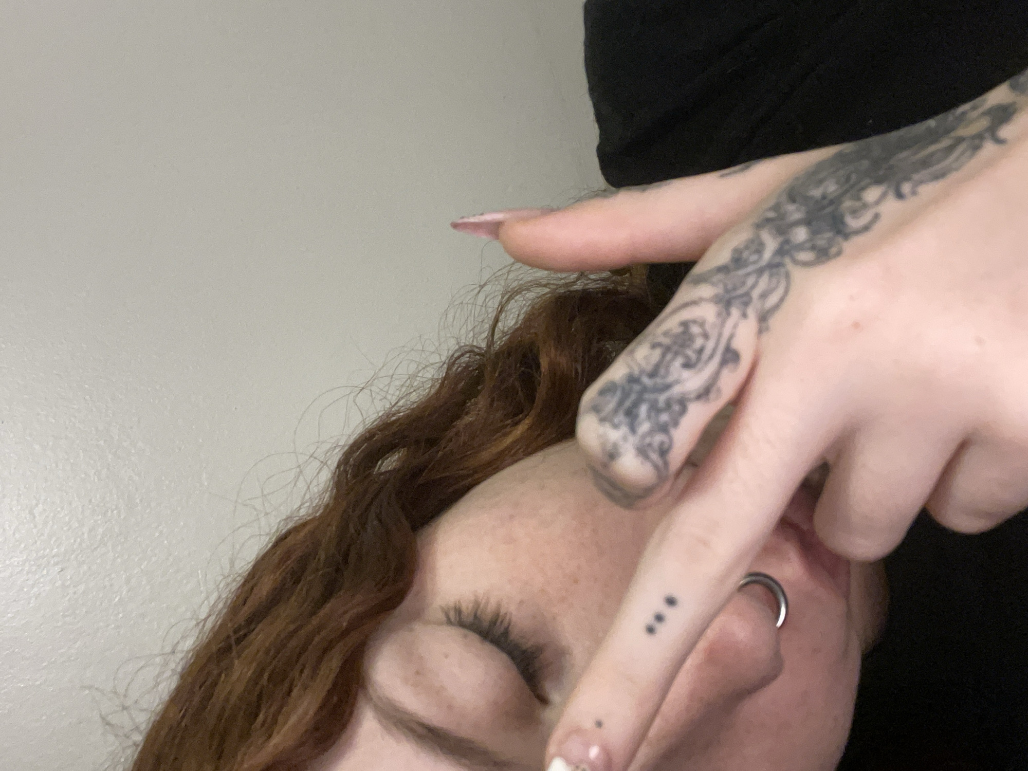A close-up shot depicts a portion of a person's face, specifically the eye, nose, and part of the forehead. The person has red-brown curly hair and freckles on their face. Their eye is closed with visible eyelashes. The nose is pierced with a silver ring. A hand is positioned over the face. The hand has detailed black tattoos on the fingers and back of the hand, as well as a row of three small dots tattooed on the index finger. The background appears to be a plain, light-colored wall, with a dark or black fabric visible behind the hand. The location of the image is not discernible as Buenos Aires, Argentina or any other location.