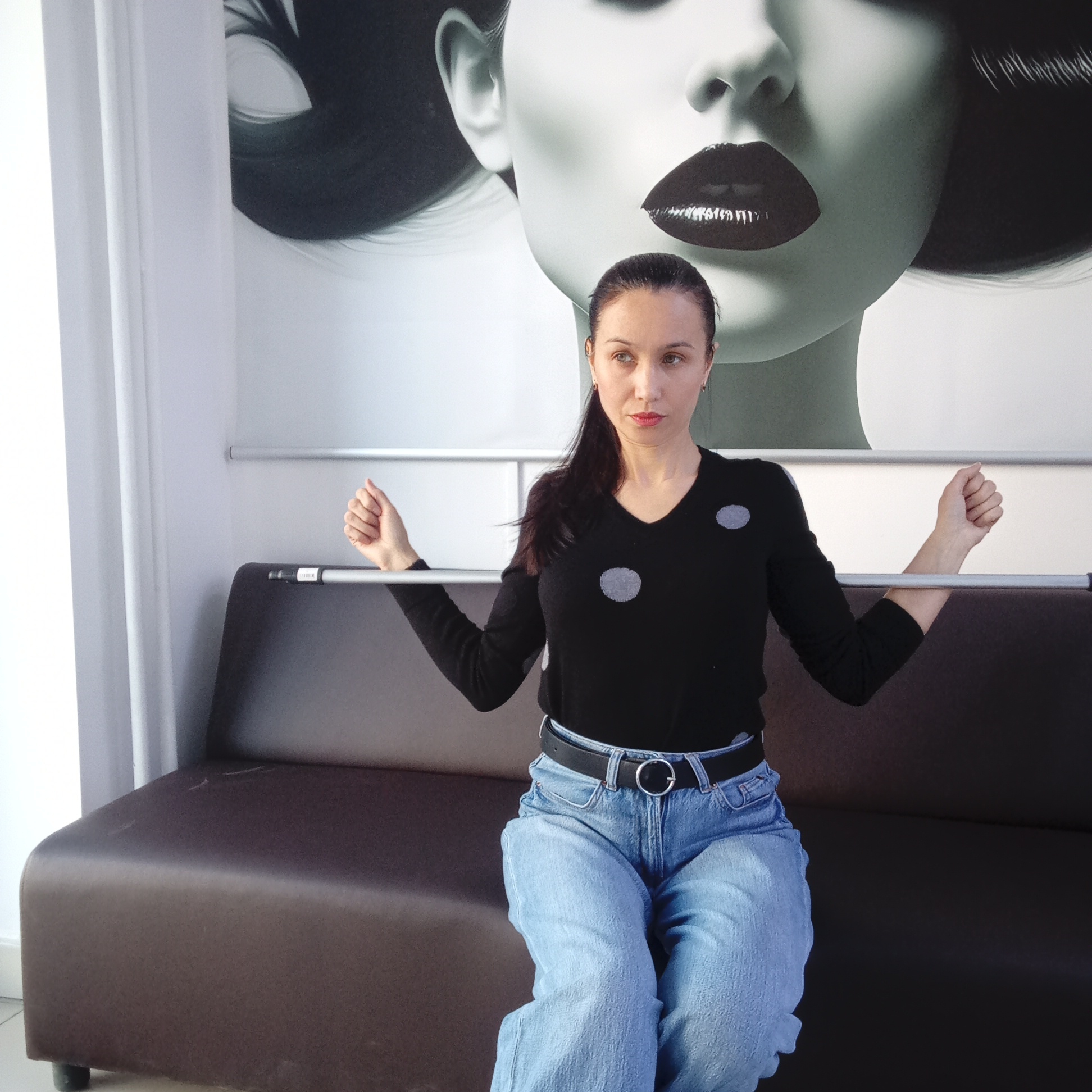 A young adult woman is seated on a dark brown sofa in an indoor setting, possibly a waiting room or salon in Izhevsk, Russia. She has light skin, dark hair pulled back in a ponytail, and a neutral, slightly serious expression as she looks to her left. She is wearing a black V-neck sweater with three grey polka dots, light blue loose-fitting jeans, and a black belt with a silver round buckle.
The woman is holding onto a horizontal silver metal bar with both hands, positioned behind her back at elbow height. Her right hand is clenched in a fist, while her left hand is more open, with a small, unreadable white label visible on the bar near it. Above this bar, a second identical silver bar is mounted on the wall.
Behind the woman and the sofa, a large, stylized graphic illustration of a woman's face dominates the white wall. The graphic features a woman with dark hair, dark, glossy lips, and a prominent nose and chin, rendered in shades of black, grey, and white. Bright, ambient lighting from the left side of the frame suggests it is daytime. A white vertical column or curtain is visible on the far left.
